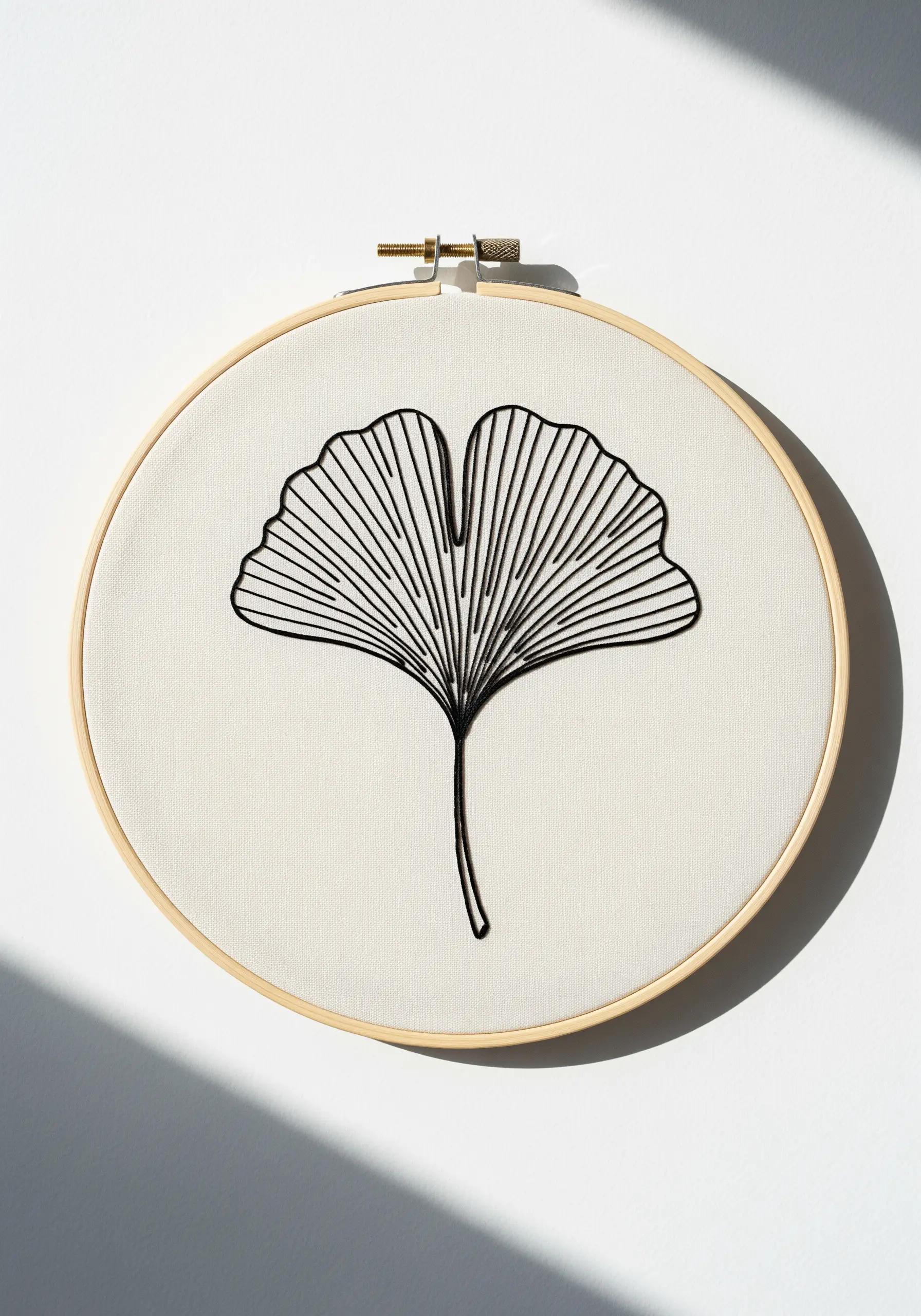 Minimalist black thread embroidery of a ginkgo leaf with detailed veins on white fabric.
