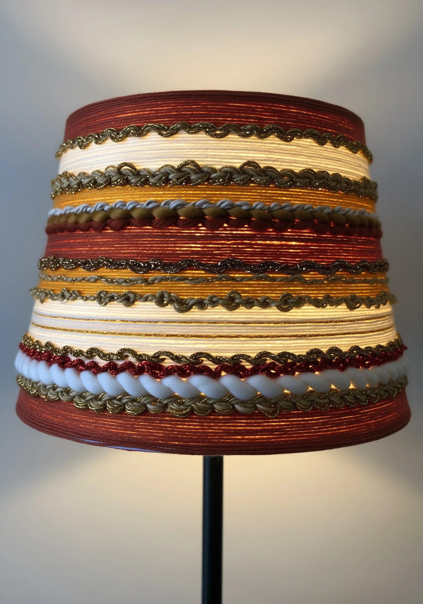 A lampshade decorated with horizontal bands of wrapped yarns and decorative braids in warm, earthy tones.