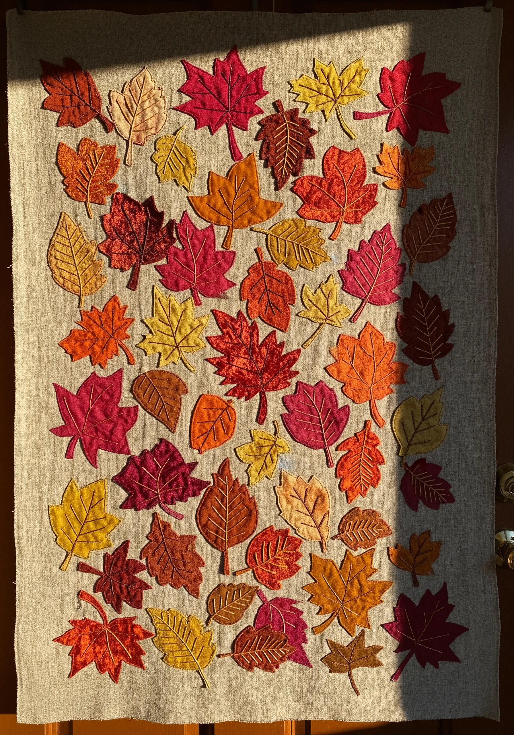 A wall hanging covered in scattered, appliquéd autumn leaves in shades of red, orange, and yellow