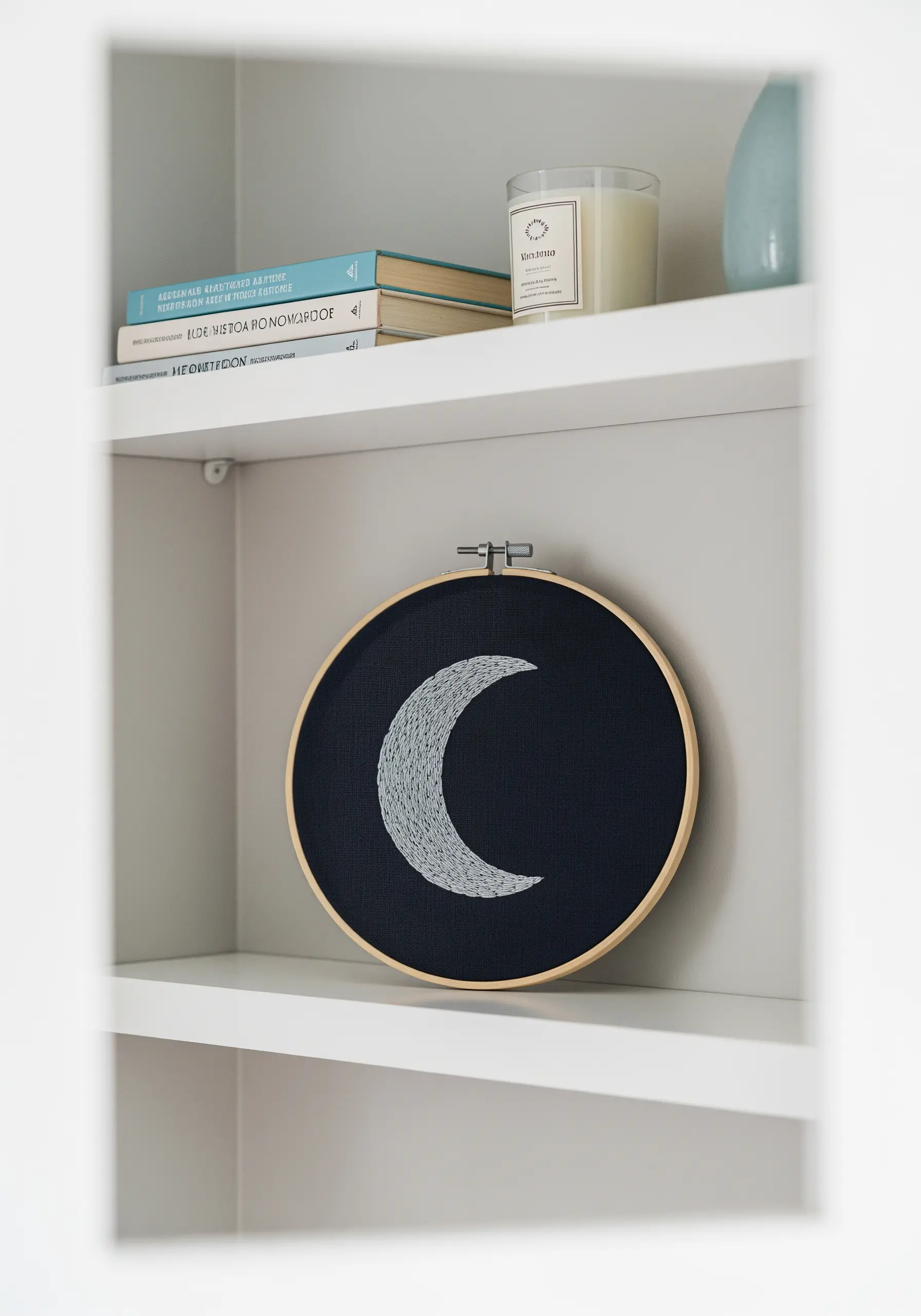 An embroidery hoop with a textured crescent moon stitched in a white-to-grey gradient on black fabric.