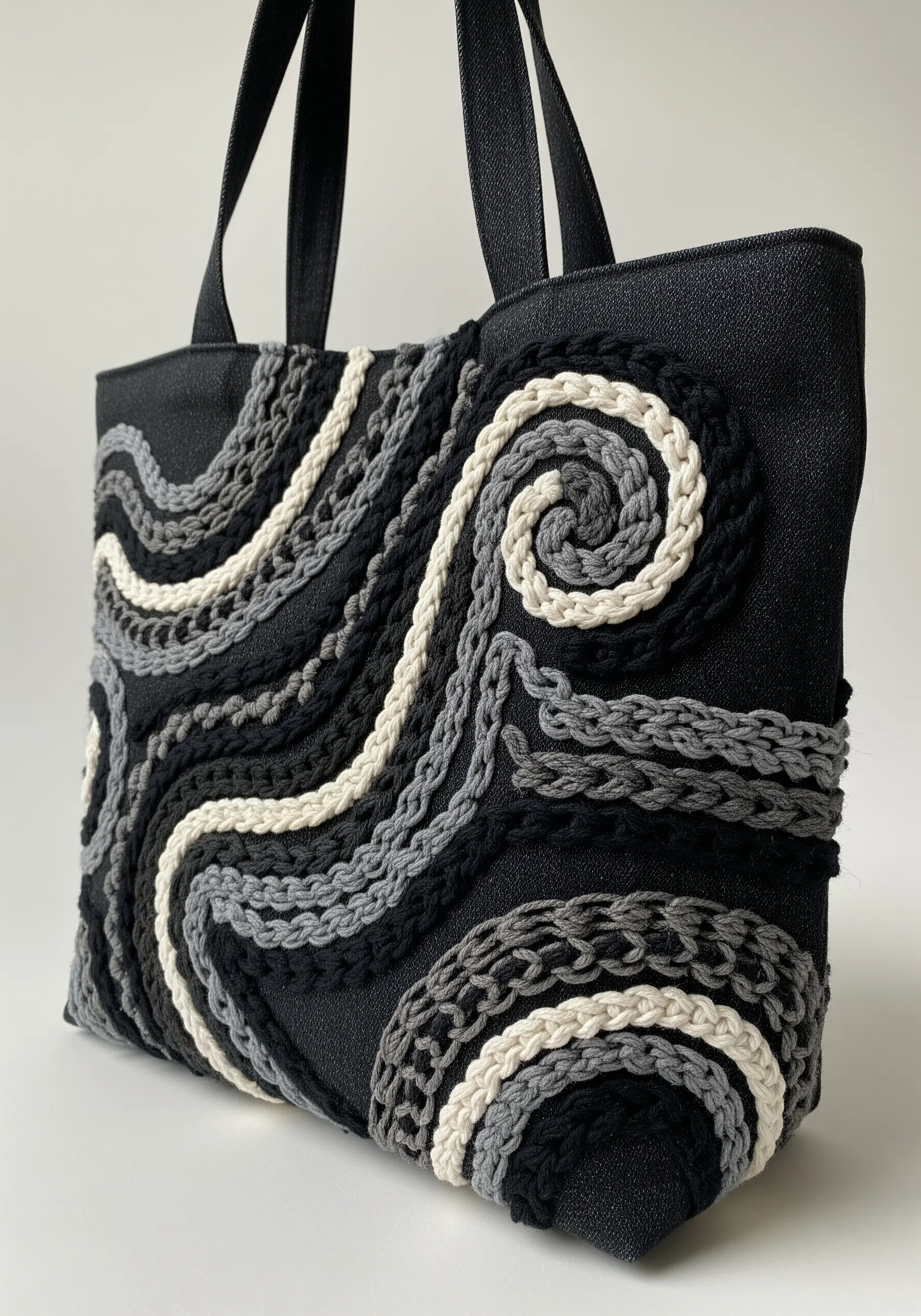 3D monochrome swirls in black, grey, and white yarn on a dark denim tote bag.
