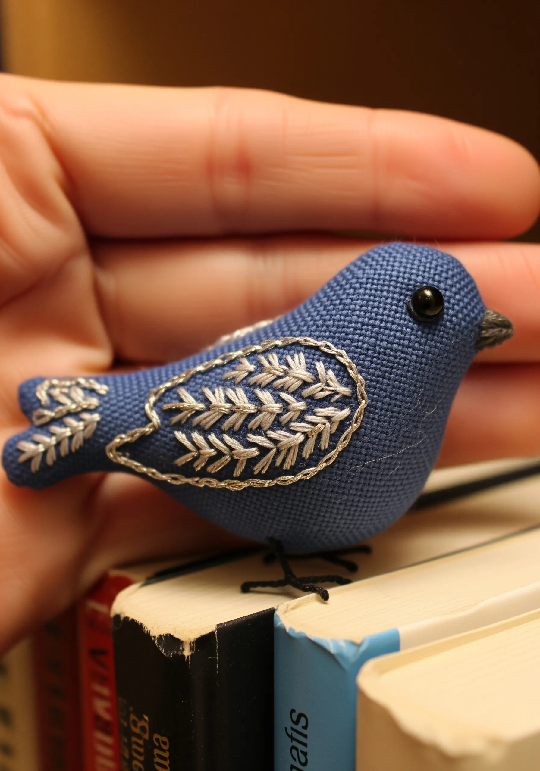 Miniature blue bird, hand-stitched with silver thread wings, perched on a book.