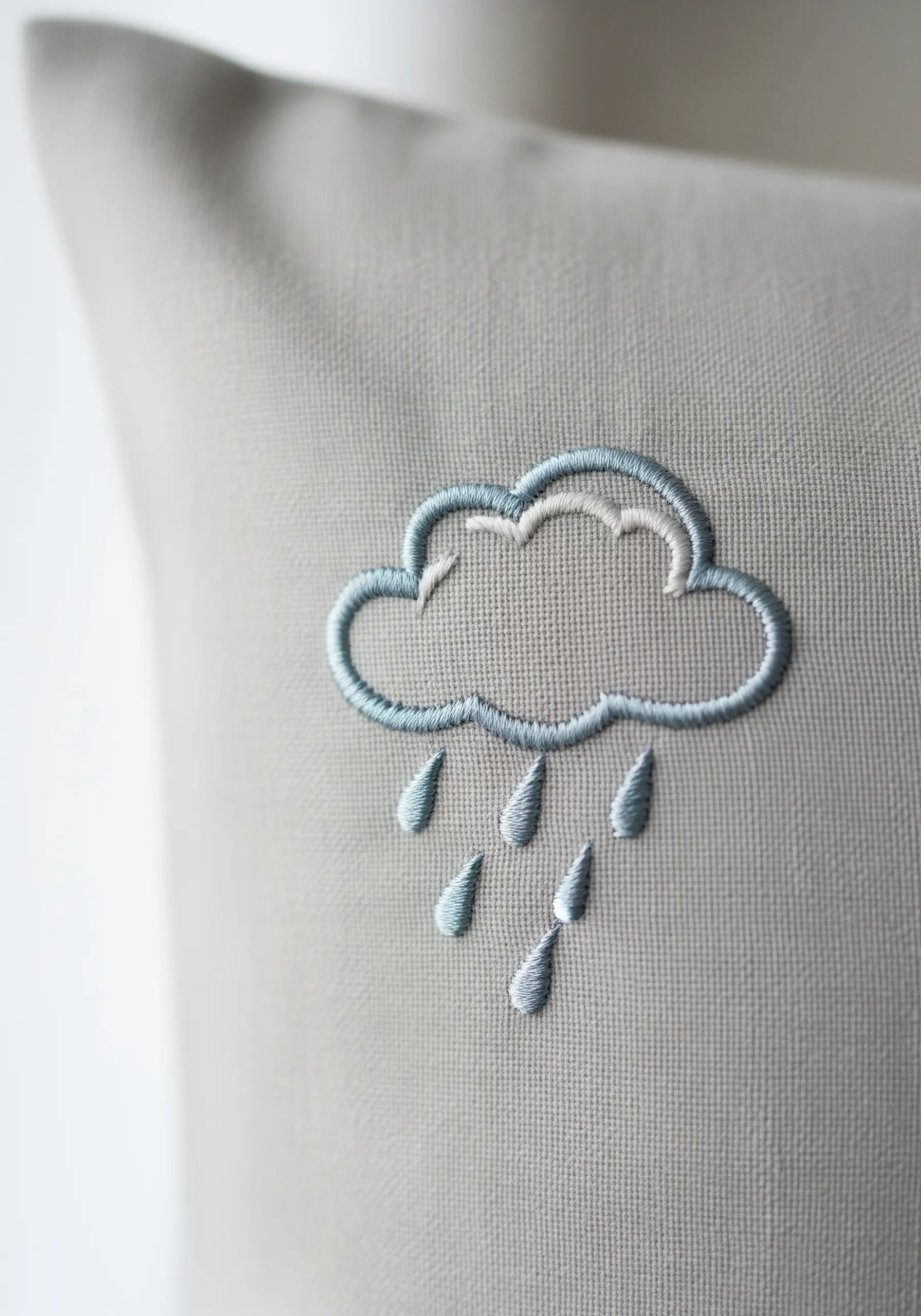 A satin-stitched cloud with raindrops in shades of blue and white on a grey pillowcase.