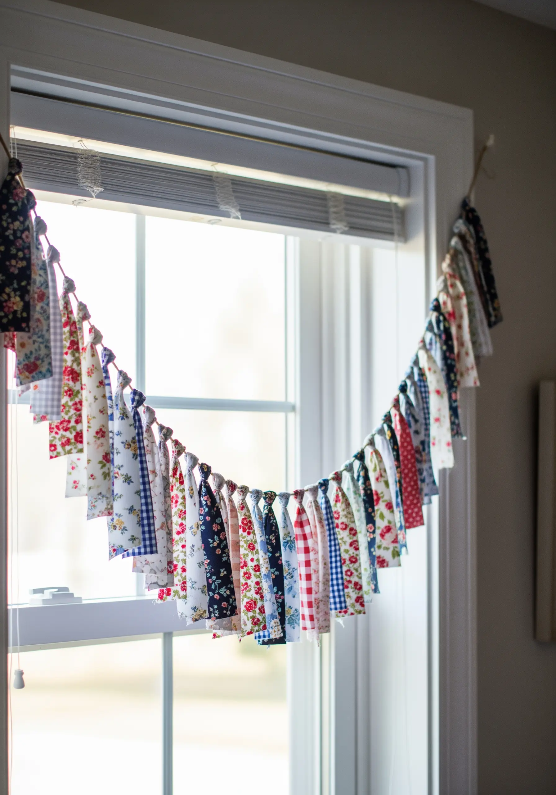 A garland made of tied fabric scraps in various floral and gingham patterns hanging in a window.