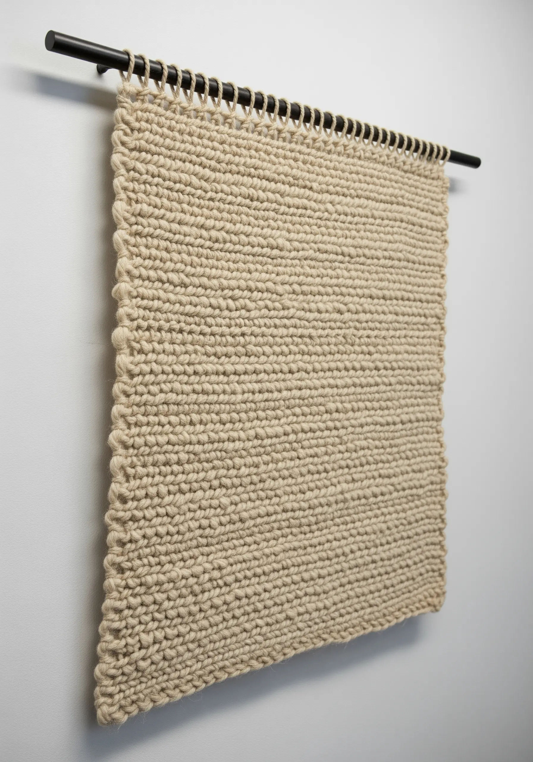 A minimalist knitted wall hanging in a neutral garter stitch, hung from a black rod.