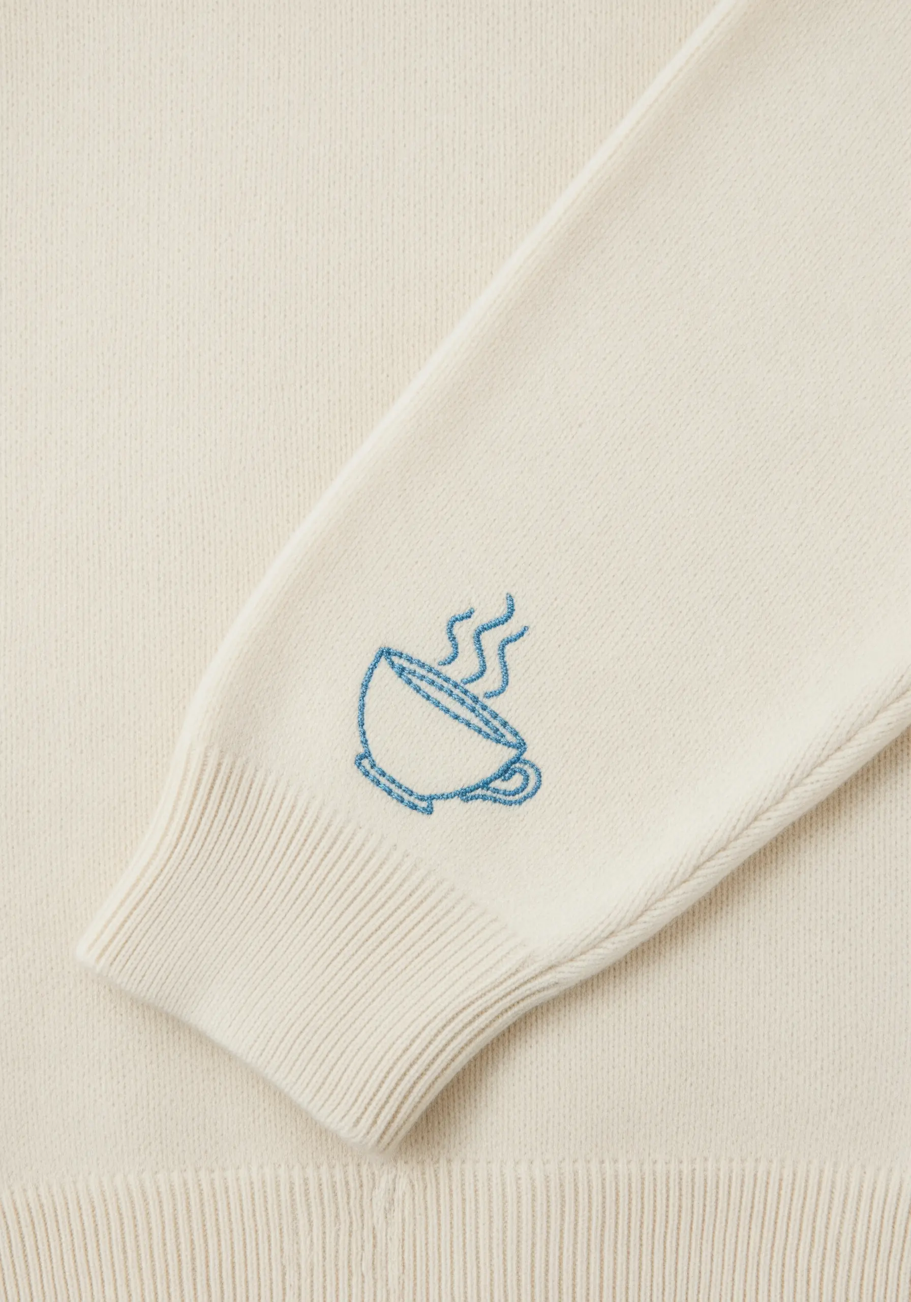 Dainty blue line art of a steaming coffee cup on a cream sweater sleeve.
