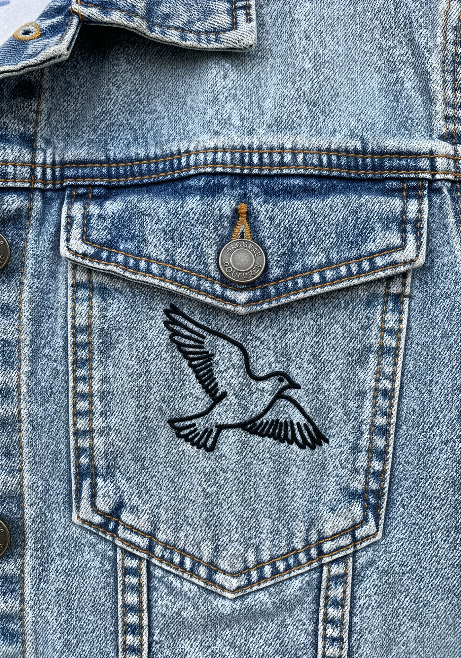 Black line art embroidery of a bird on a denim jacket pocket.