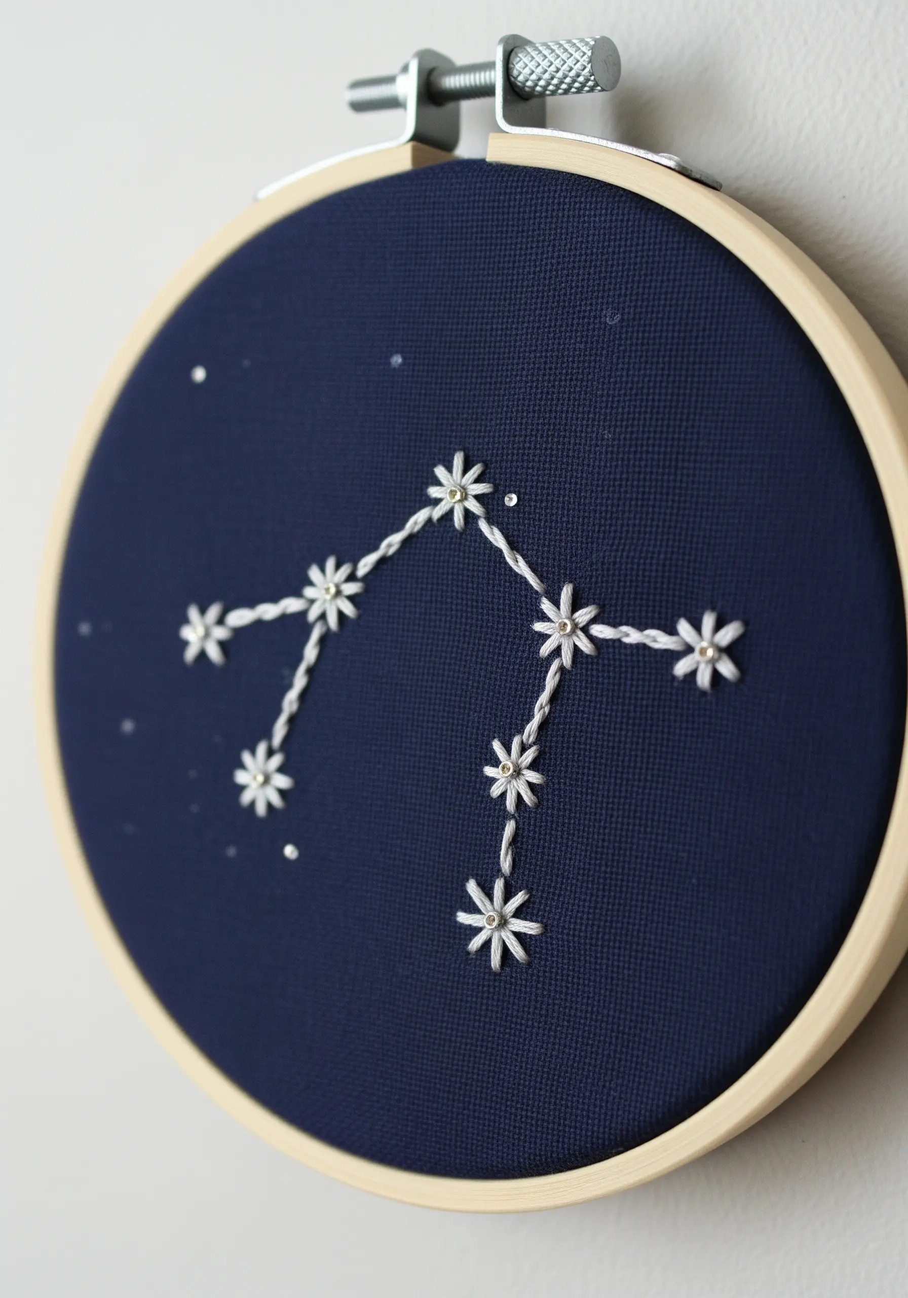An embroidery hoop with a zodiac constellation stitched in white thread on navy blue fabric.