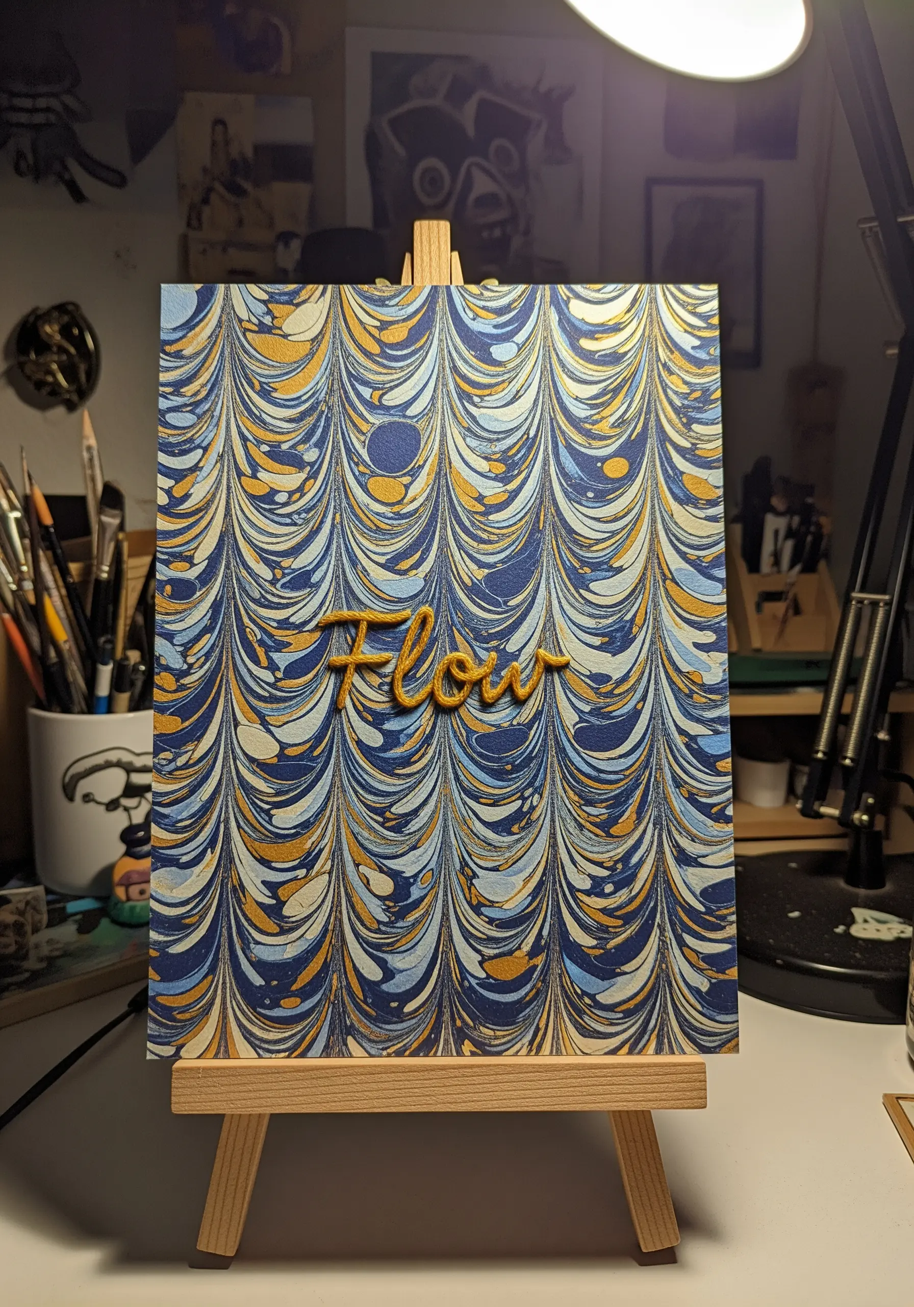 The word 'Flow' embroidered in a raised, 3D golden thread on a piece of marbled paper.
