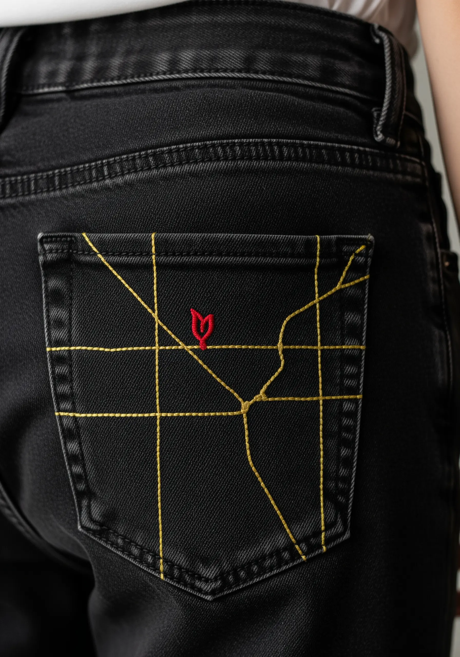 A minimalist map stitched in yellow thread on the back pocket of black jeans, with a red icon.