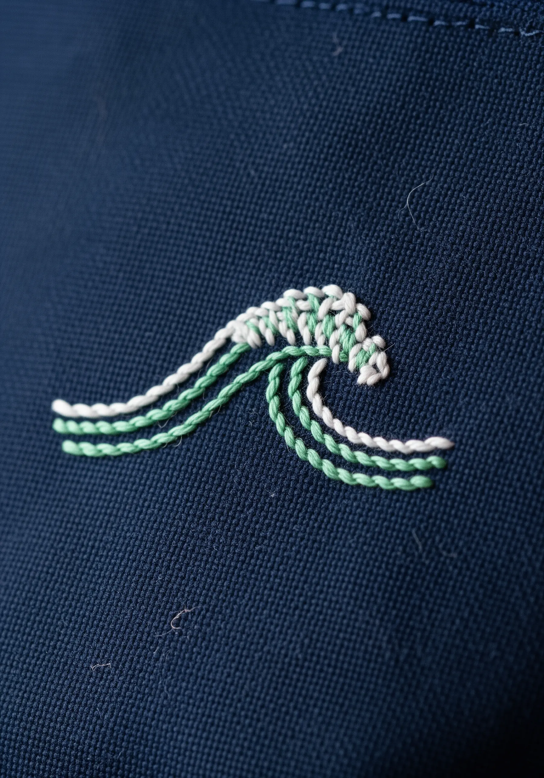A small, stylized embroidered wave in white and green on a dark blue fabric.