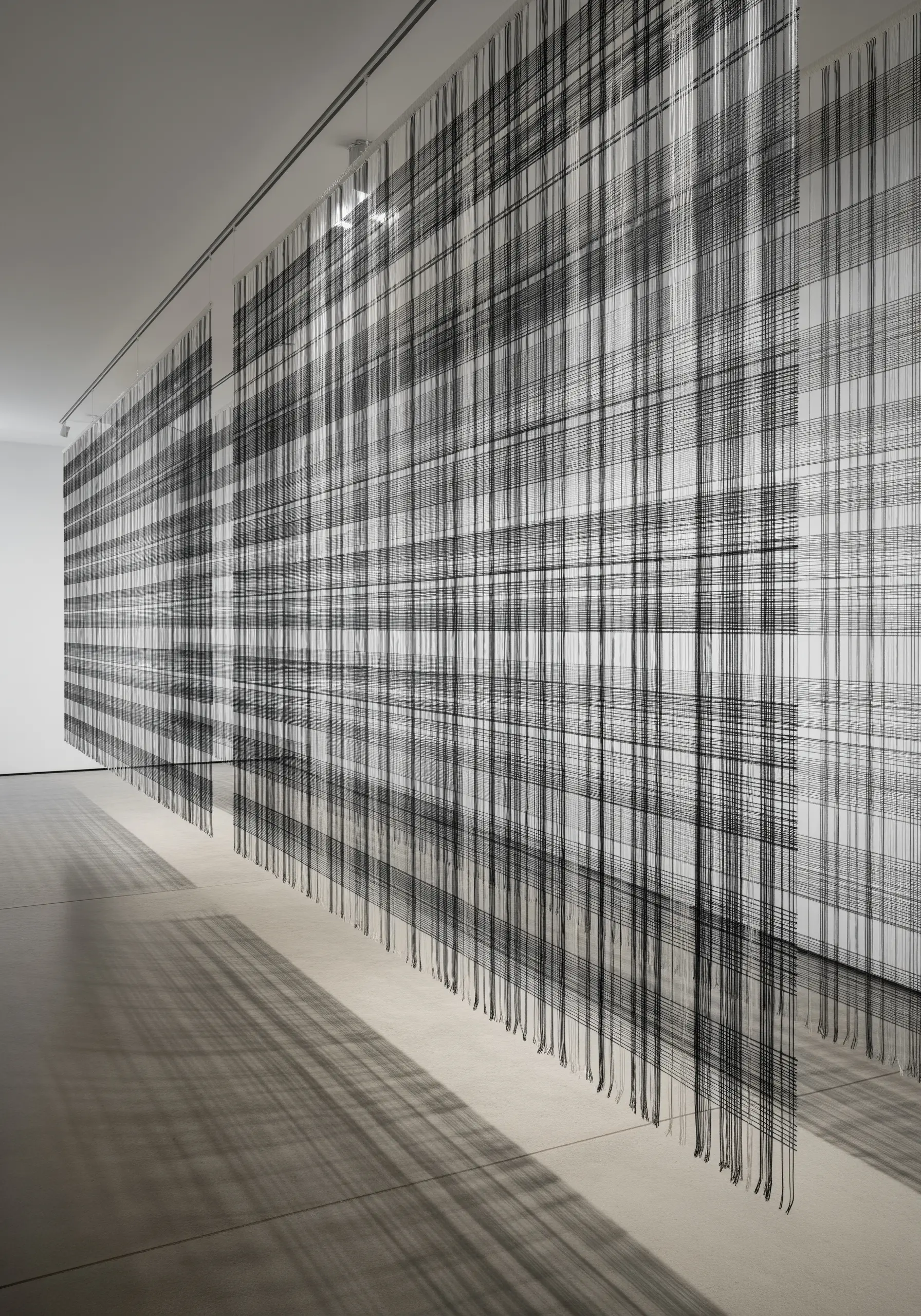 Long, parallel black threads of varying density hang from a gallery ceiling, creating a glitch effect.