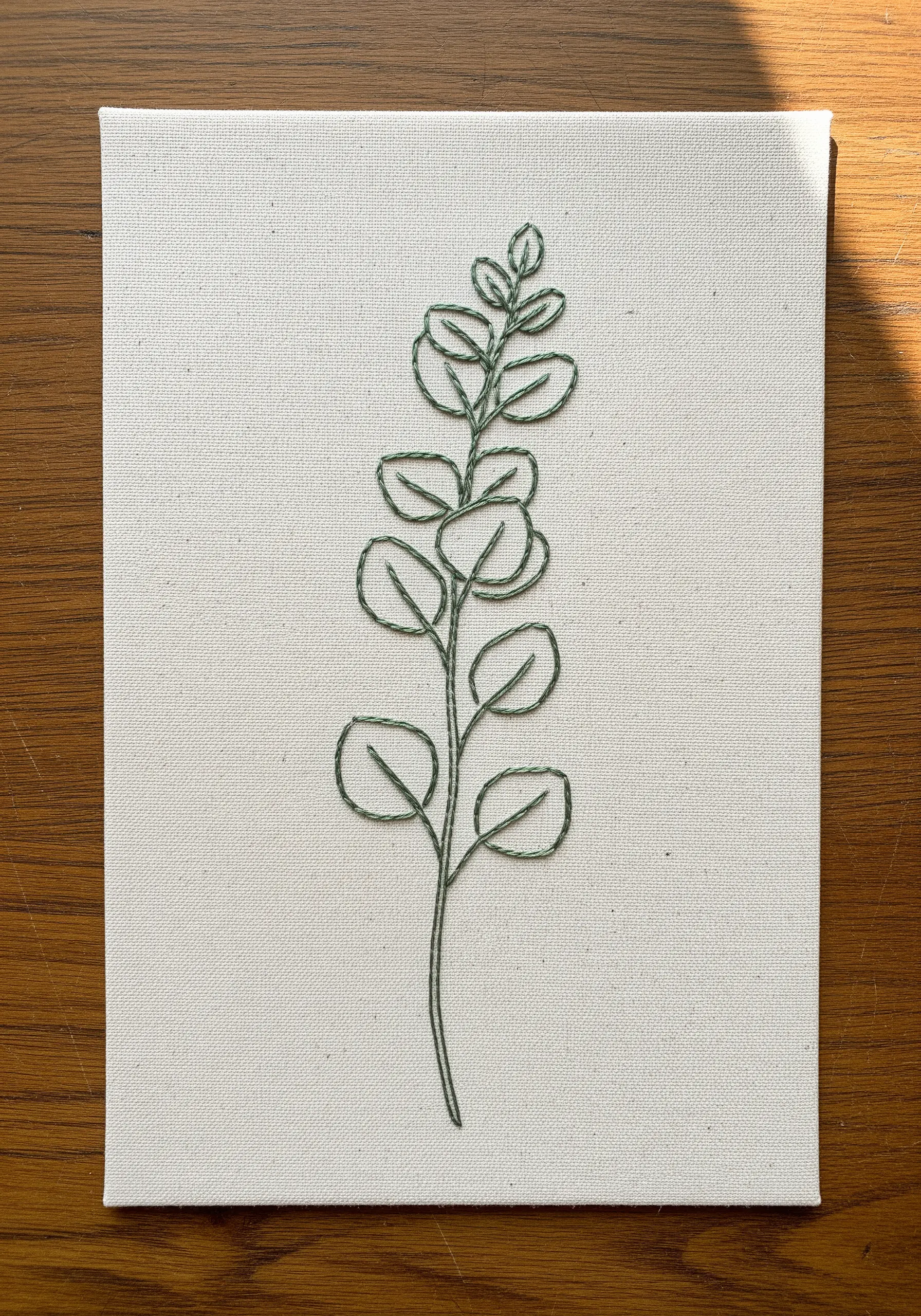 Minimalist eucalyptus branch stitched in green thread on a rectangular stretched canvas.