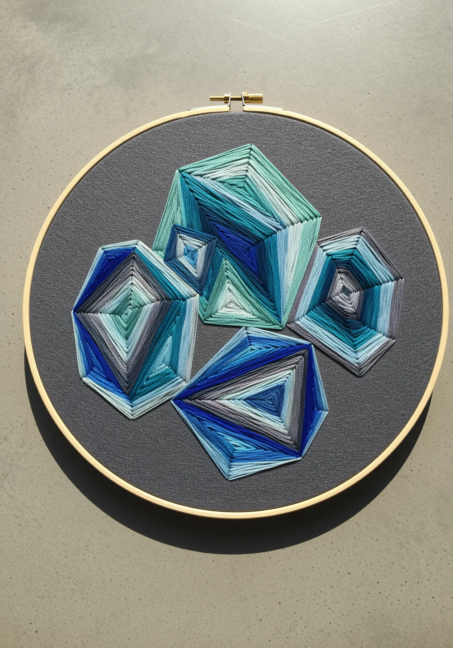 Geometric shapes in blue and green thread stitched on dark gray fabric in a hoop.