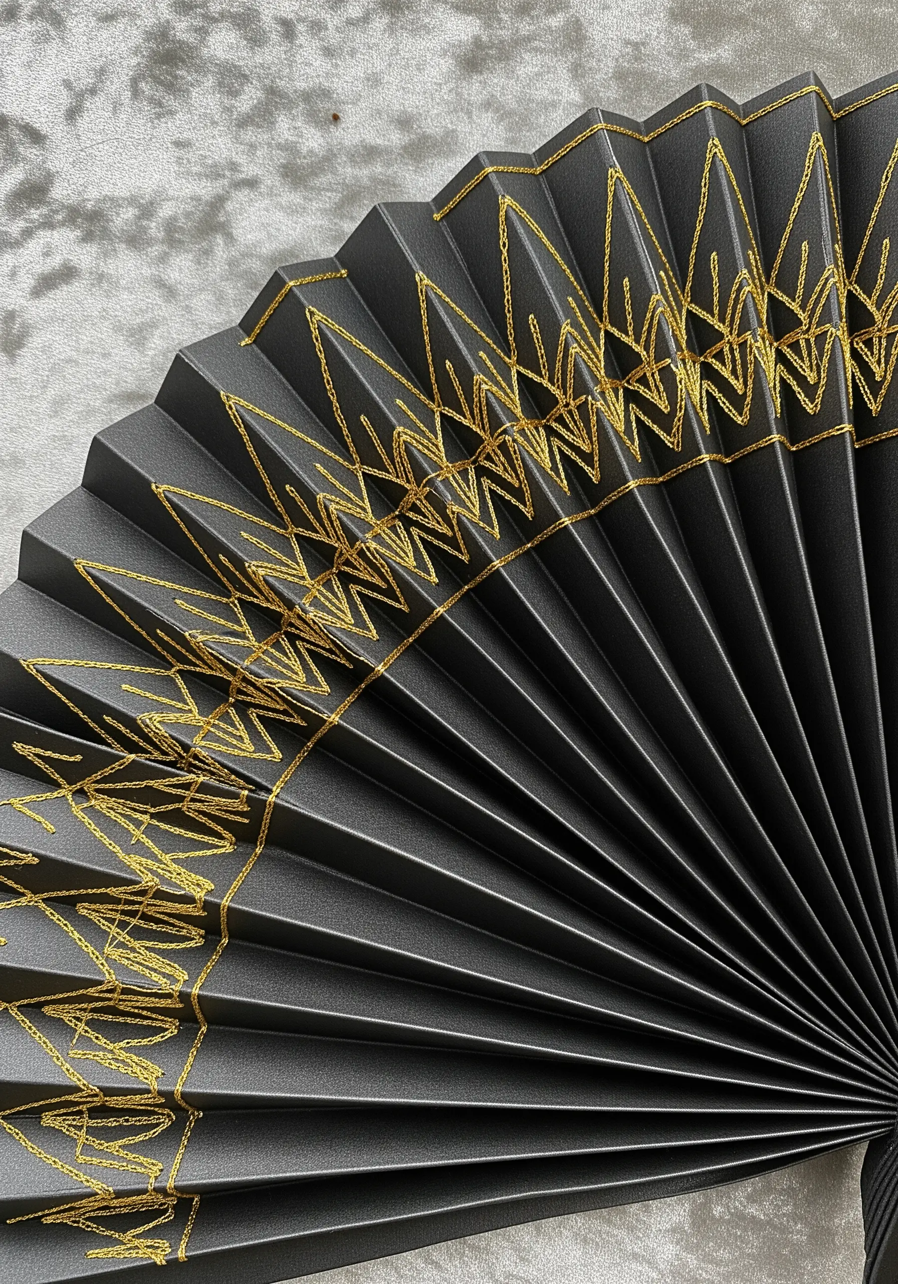 A geometric pattern stitched with gold thread on a black paper fan, creating a fractured design.