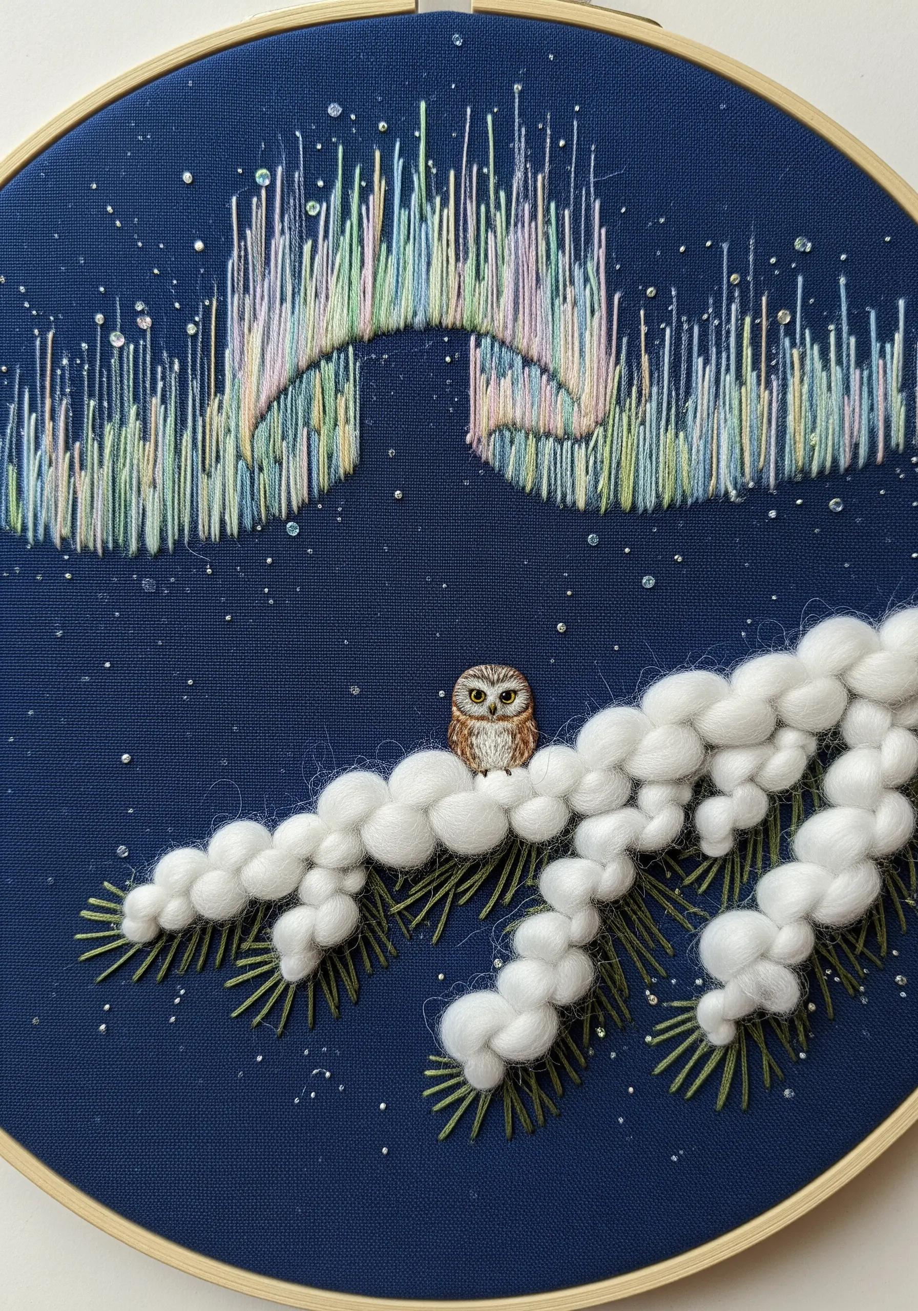 A tiny owl on a fluffy snow-covered branch, looking up at an embroidered aurora borealis in pastel colors.