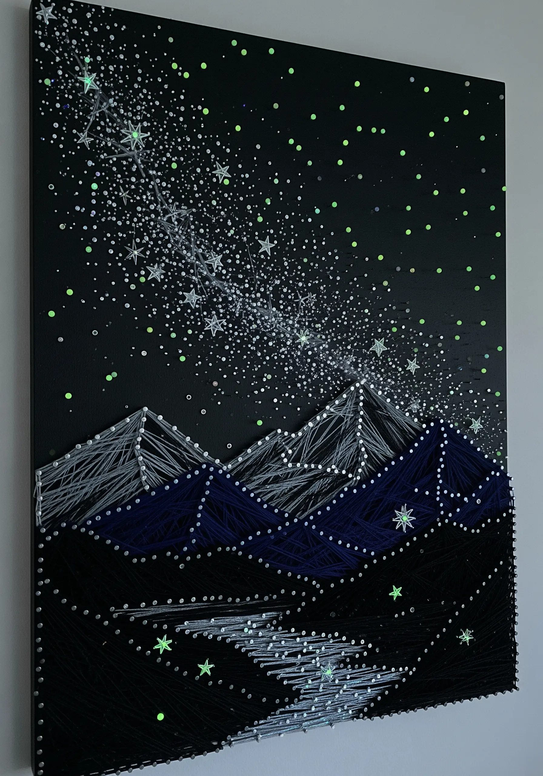 String art of a starry night sky and Milky Way over mountains, using glow-in-the-dark thread.