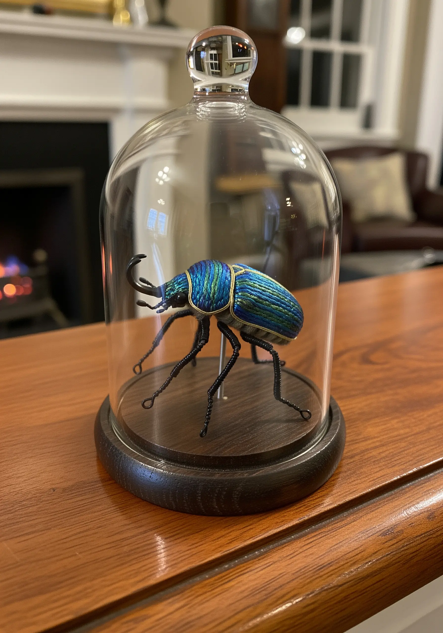 A hyper-realistic 3D embroidered beetle with iridescent thread, displayed under a glass cloche.