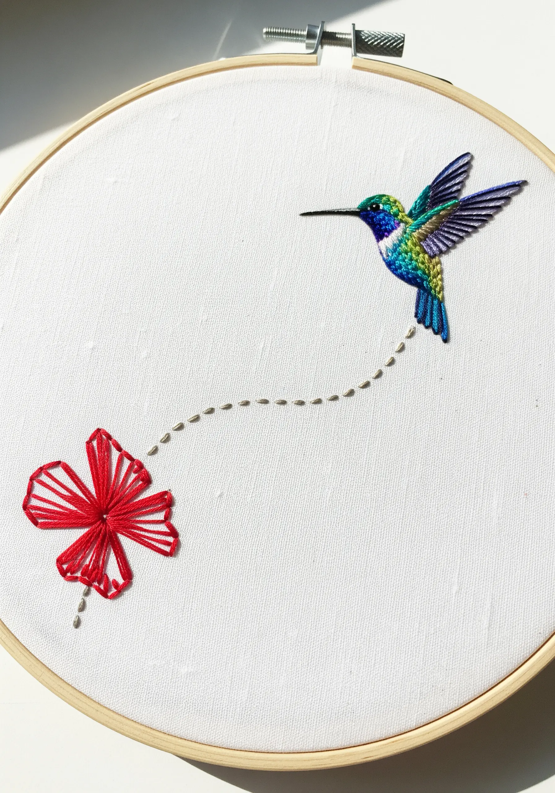 Minimalist embroidery of a hummingbird and a flower connected by a dotted line flight path.