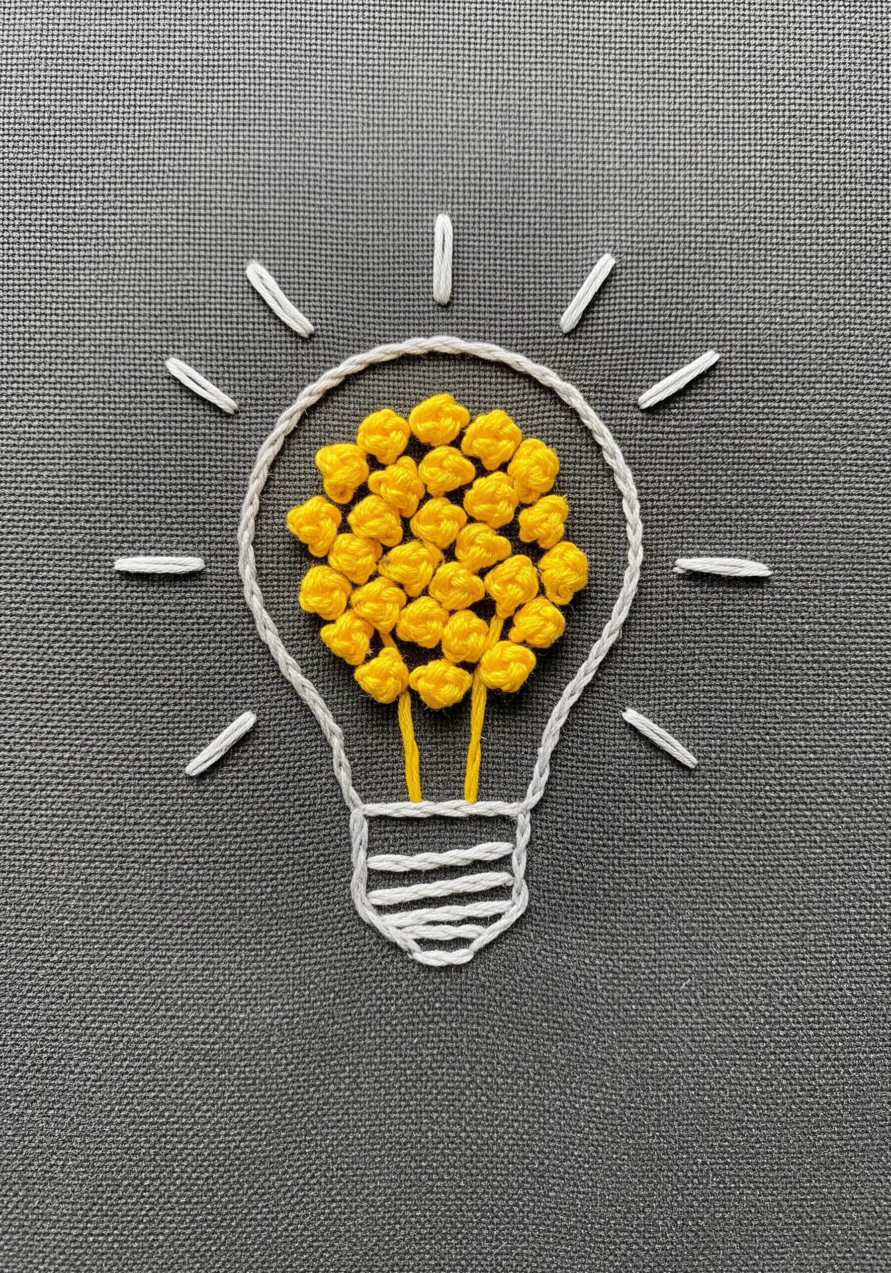 A lightbulb outlined in white thread, with the filament filled with dense yellow French knots.