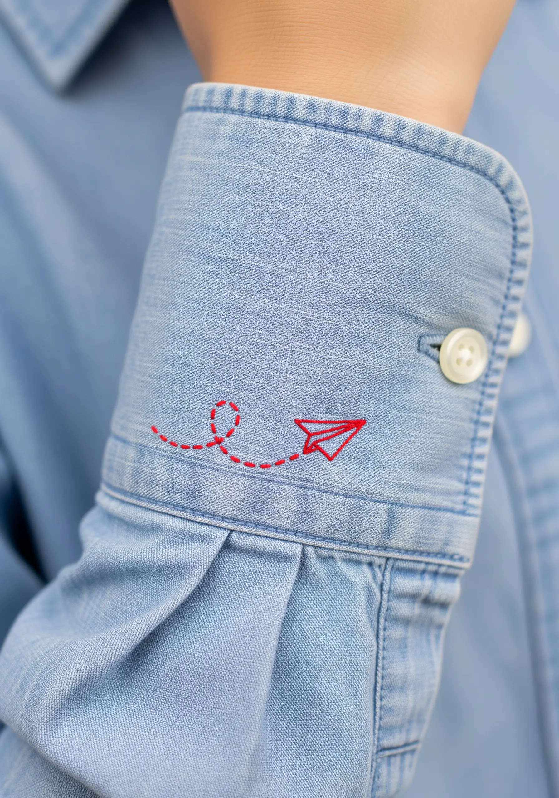 A small red paper airplane with a dashed flight path embroidered on a denim shirt cuff.
