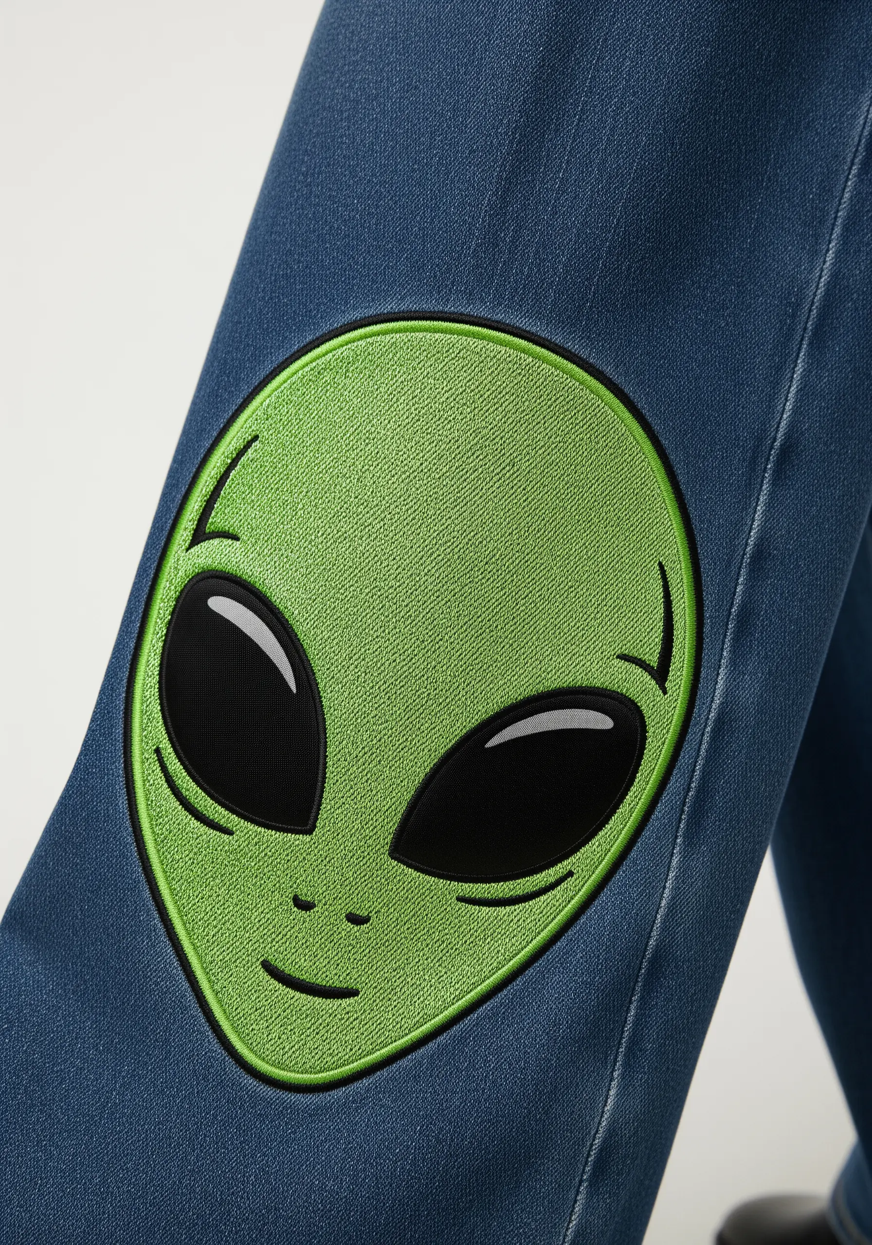 A large green alien head with glossy black eyes embroidered on a denim jean leg.