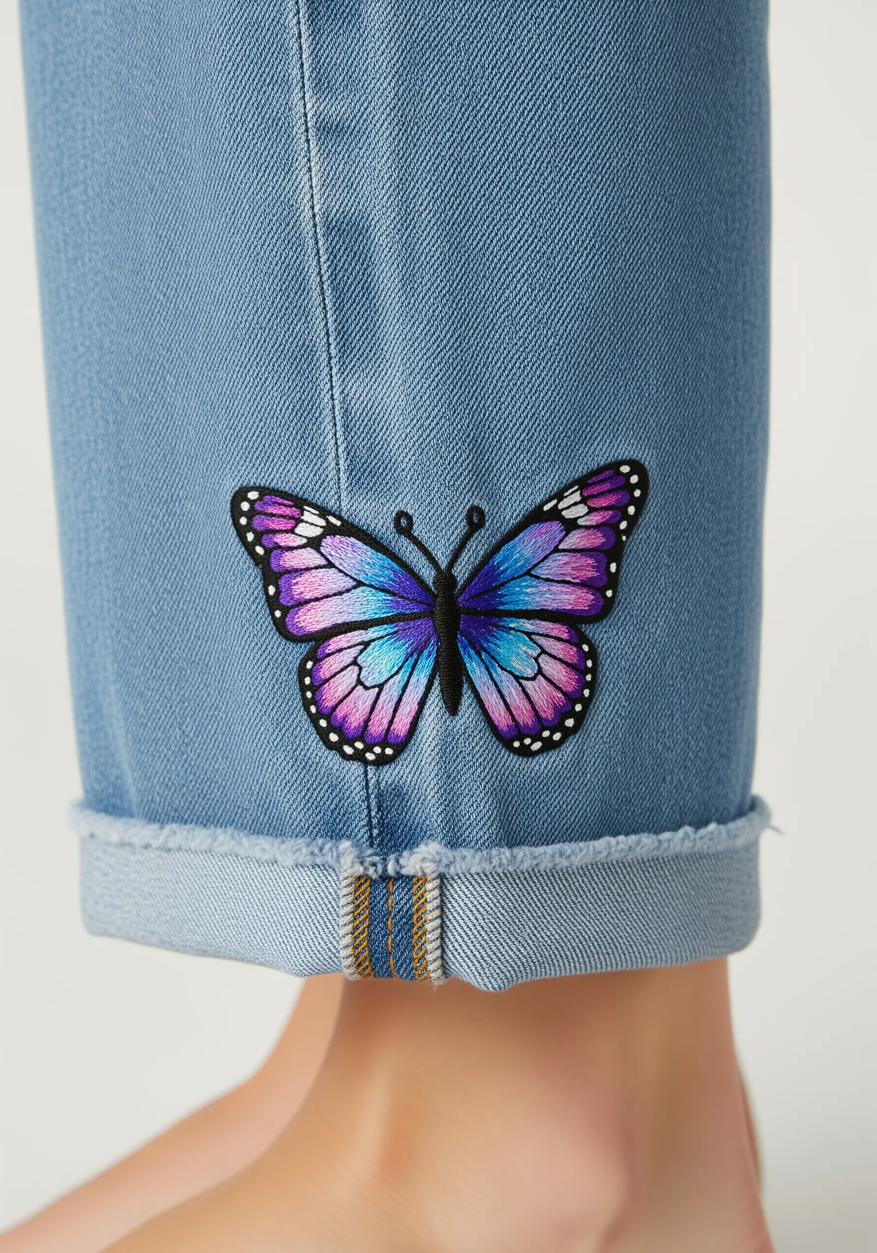 A colorful embroidered butterfly with blended purple and blue wings on a jean cuff.