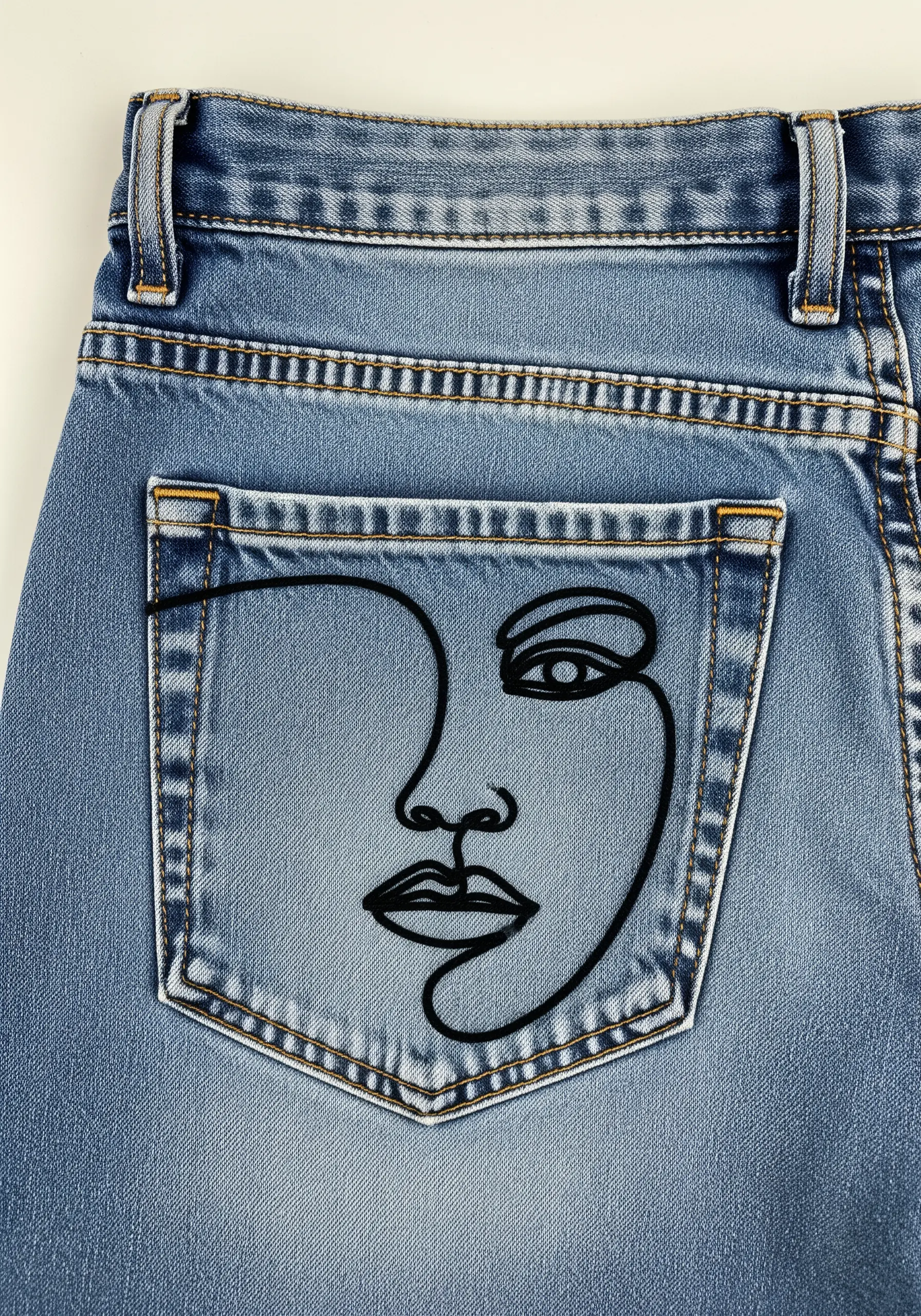 An abstract face drawn with a single black line embroidered onto a jean pocket.