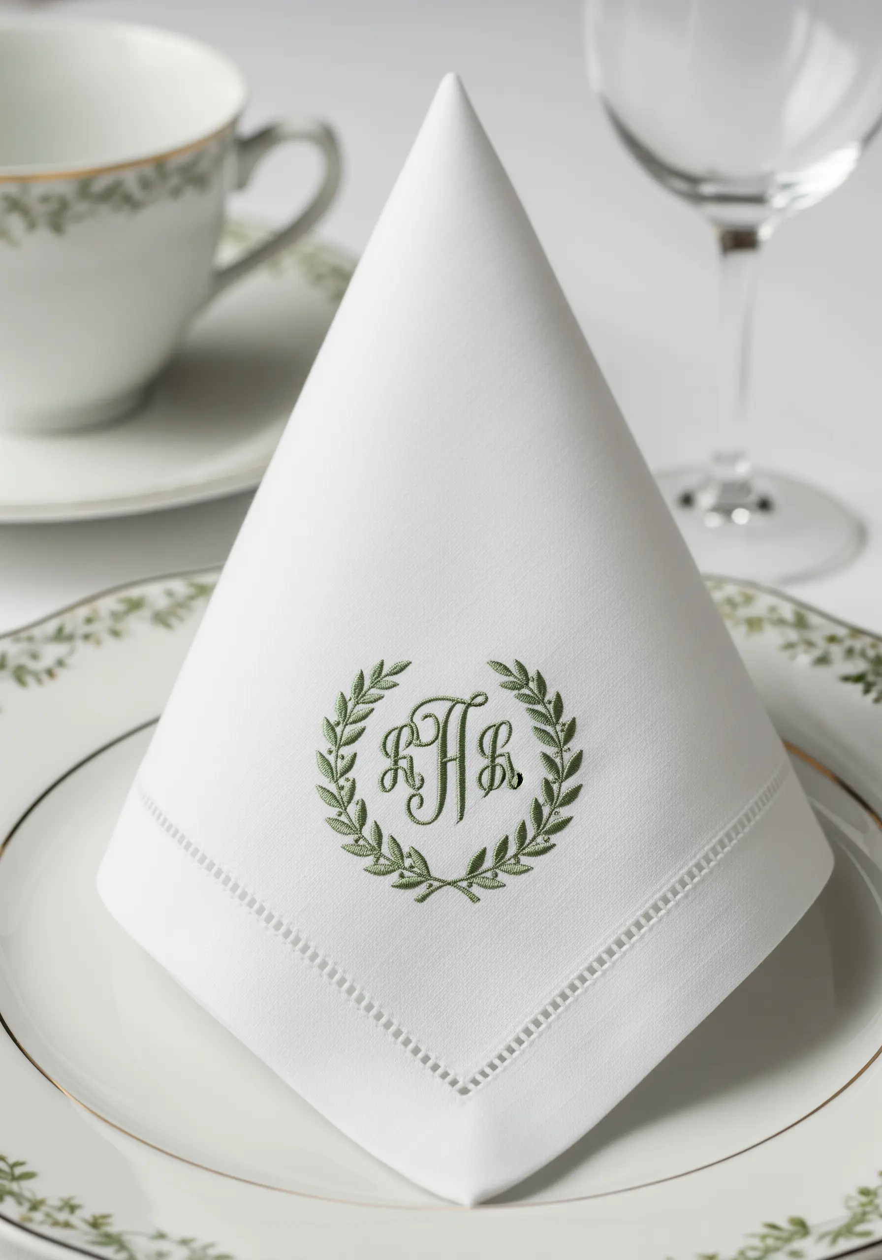 A crisp white linen napkin embroidered with a green laurel wreath surrounding a classic monogram.
