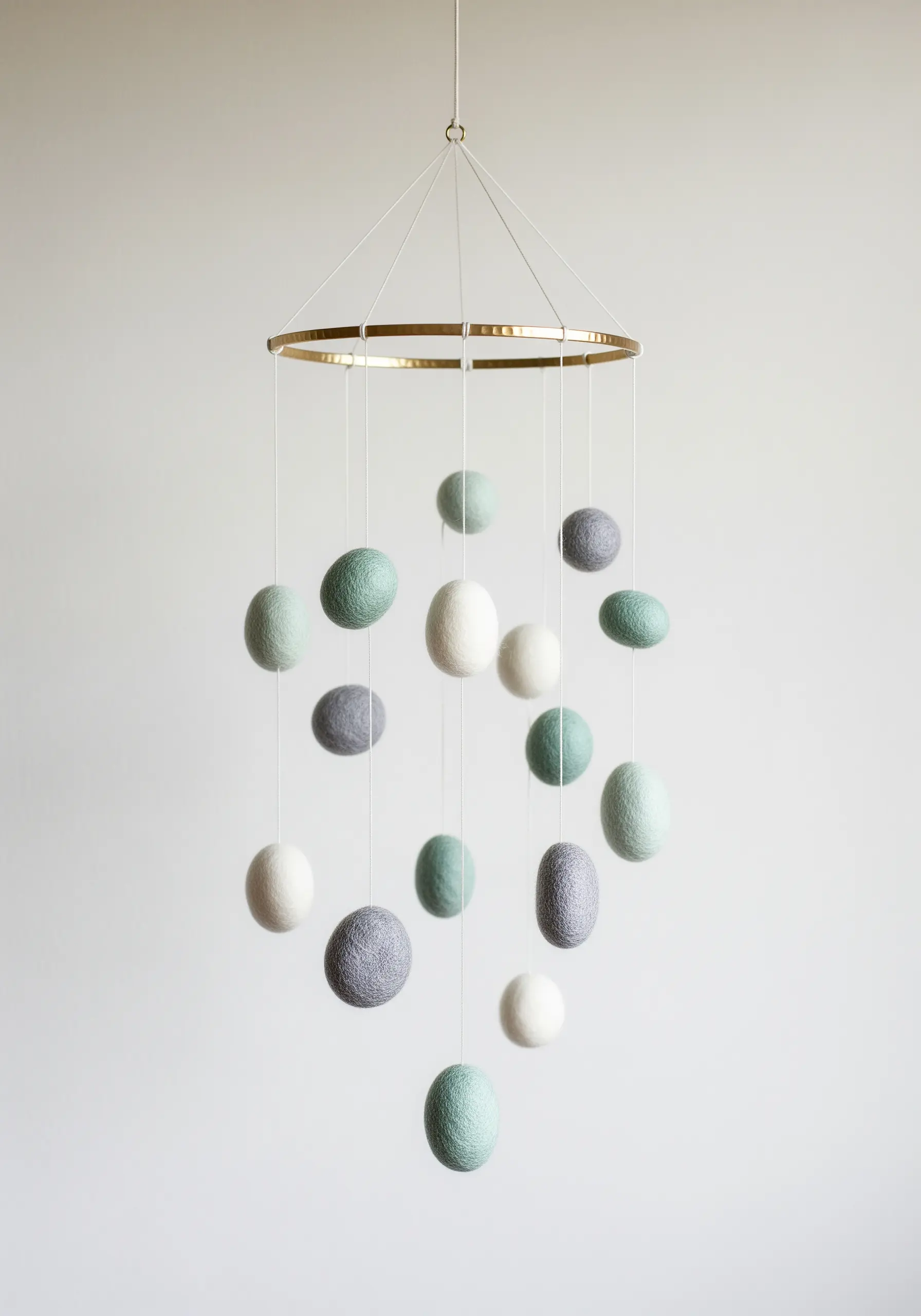 A mobile with wool felt balls in white, grey, and sage green hanging from a brass hoop.