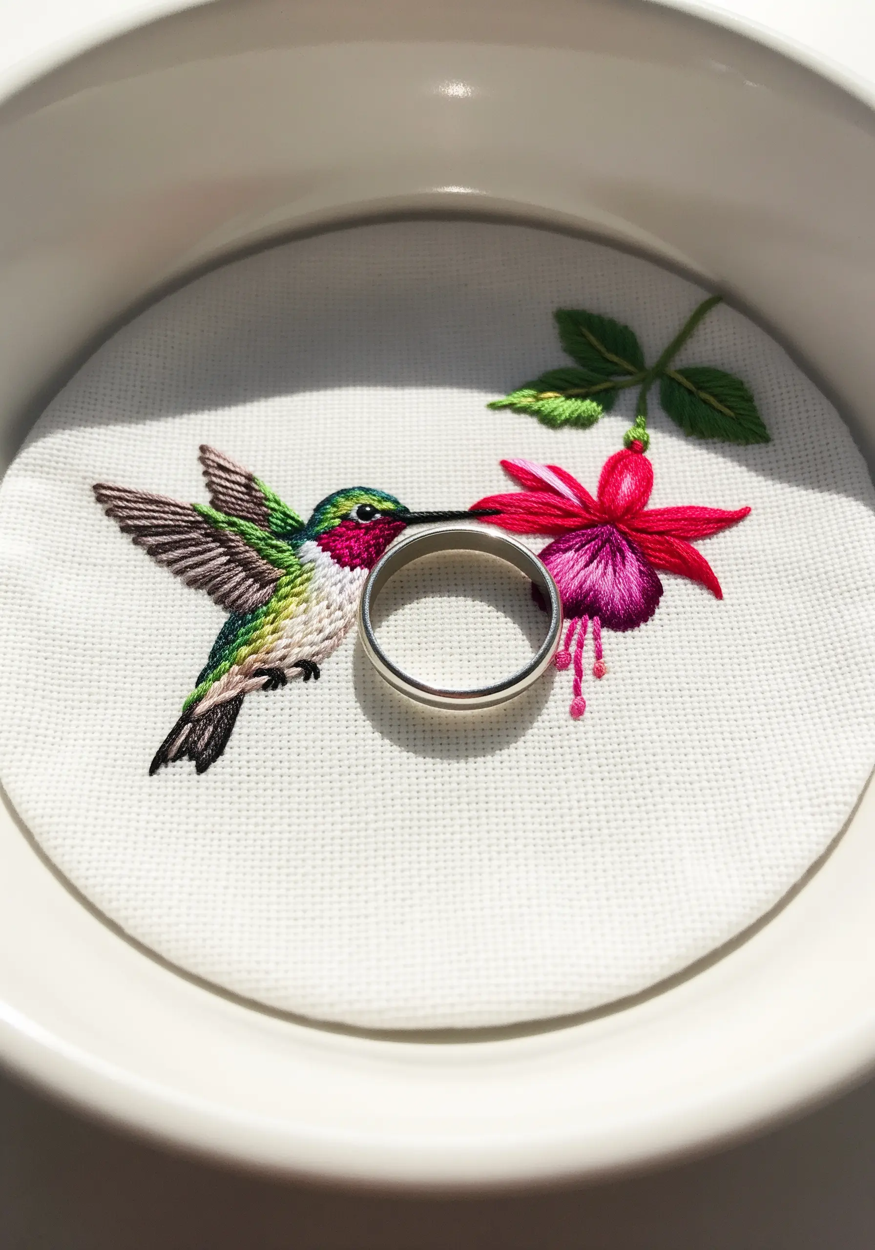 A tiny, detailed embroidery of a hummingbird and a fuchsia flower mounted in a white ring dish.