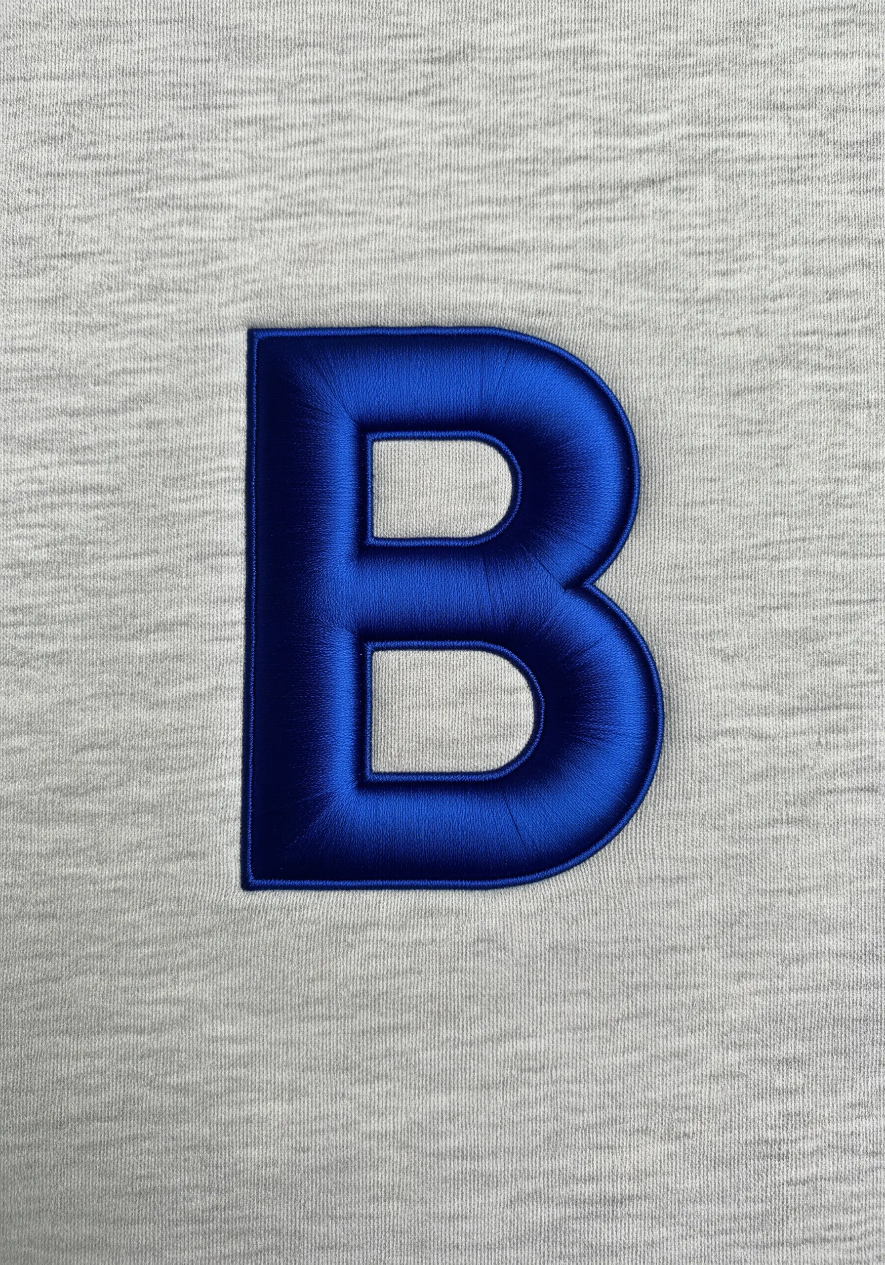 Raised, 3D satin stitch monogram 'B' in royal blue on a grey knit fabric.