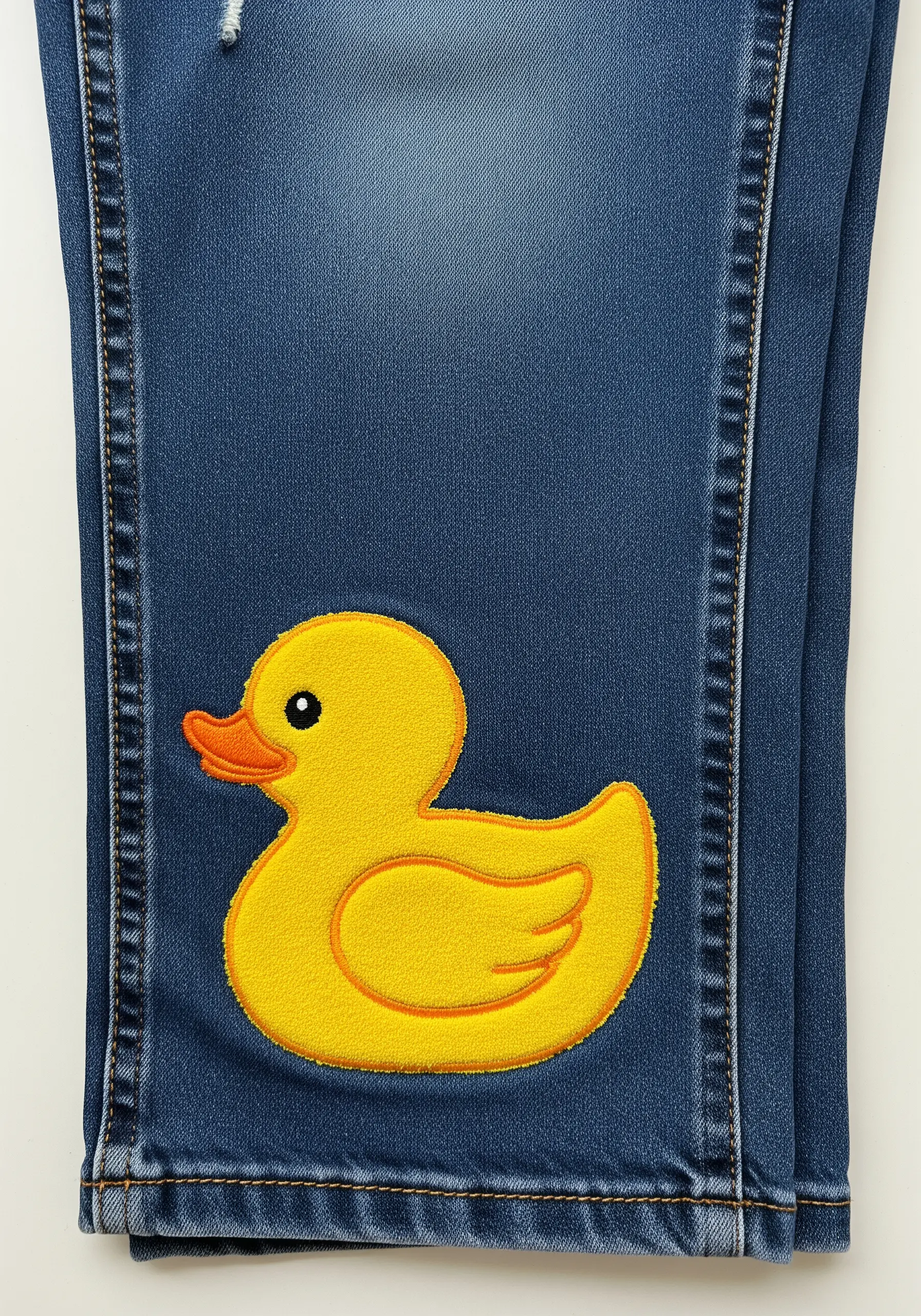 A bright yellow rubber duck with an orange outline embroidered on a dark denim leg.