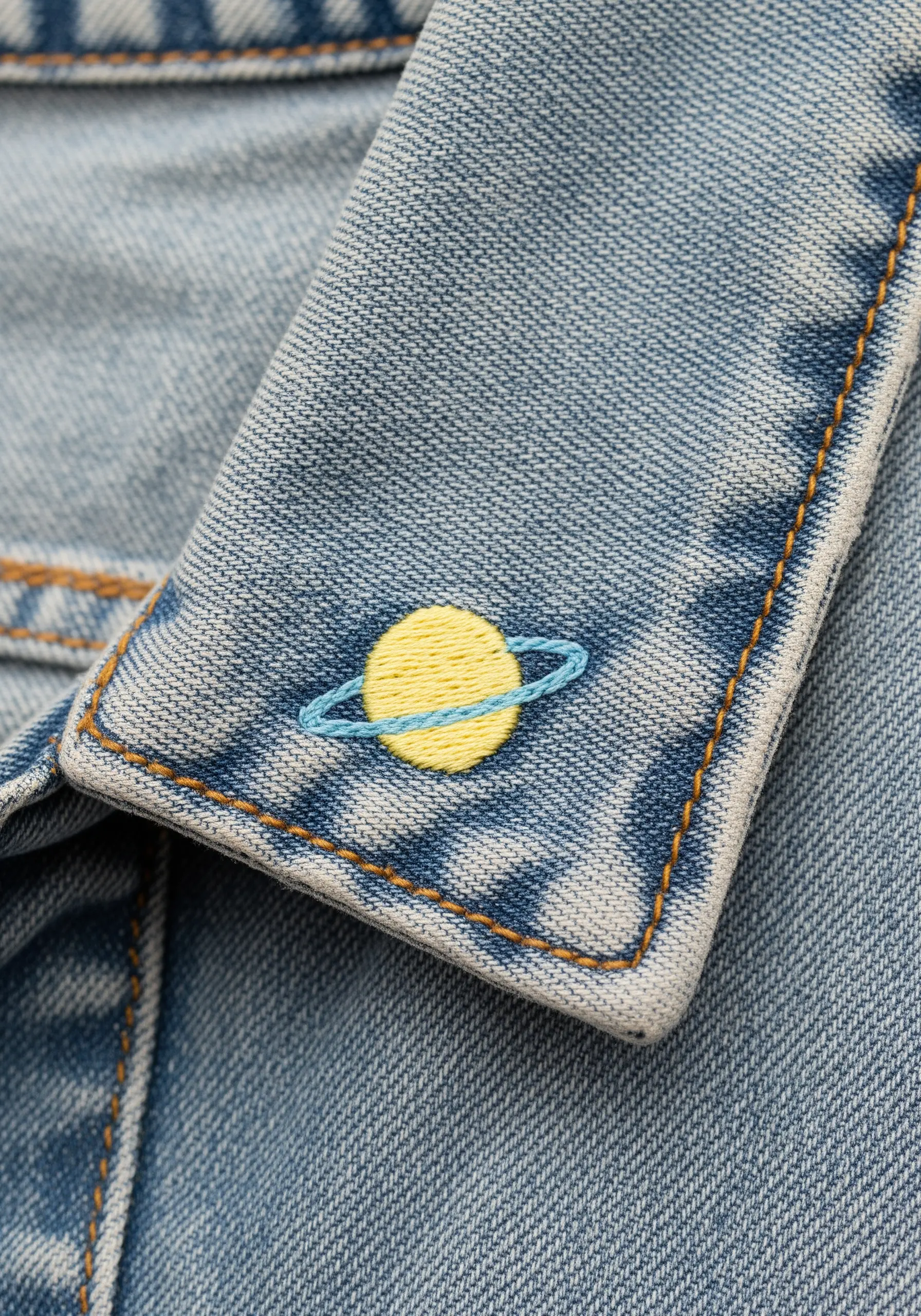 A small embroidered planet with rings, stitched on the collar point of a denim jacket