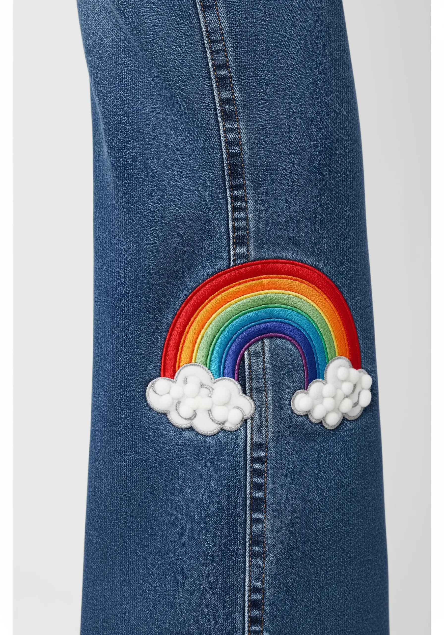 An embroidered rainbow with puffy, textured white clouds on a dark denim jean leg.