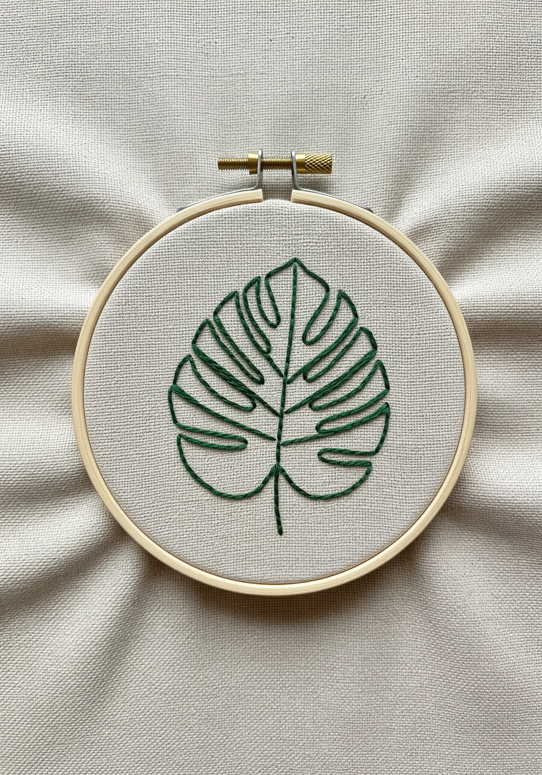 Minimalist green thread monstera leaf outline on neutral fabric.
