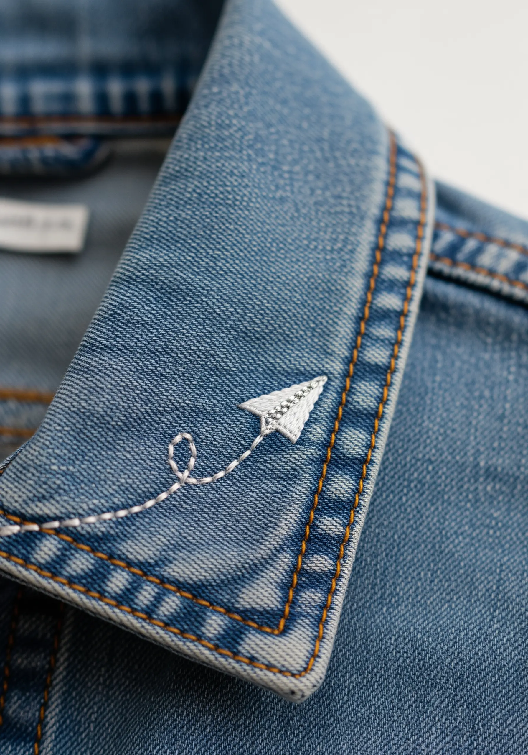 Tiny embroidered paper airplane with a looped trail on a denim jacket collar