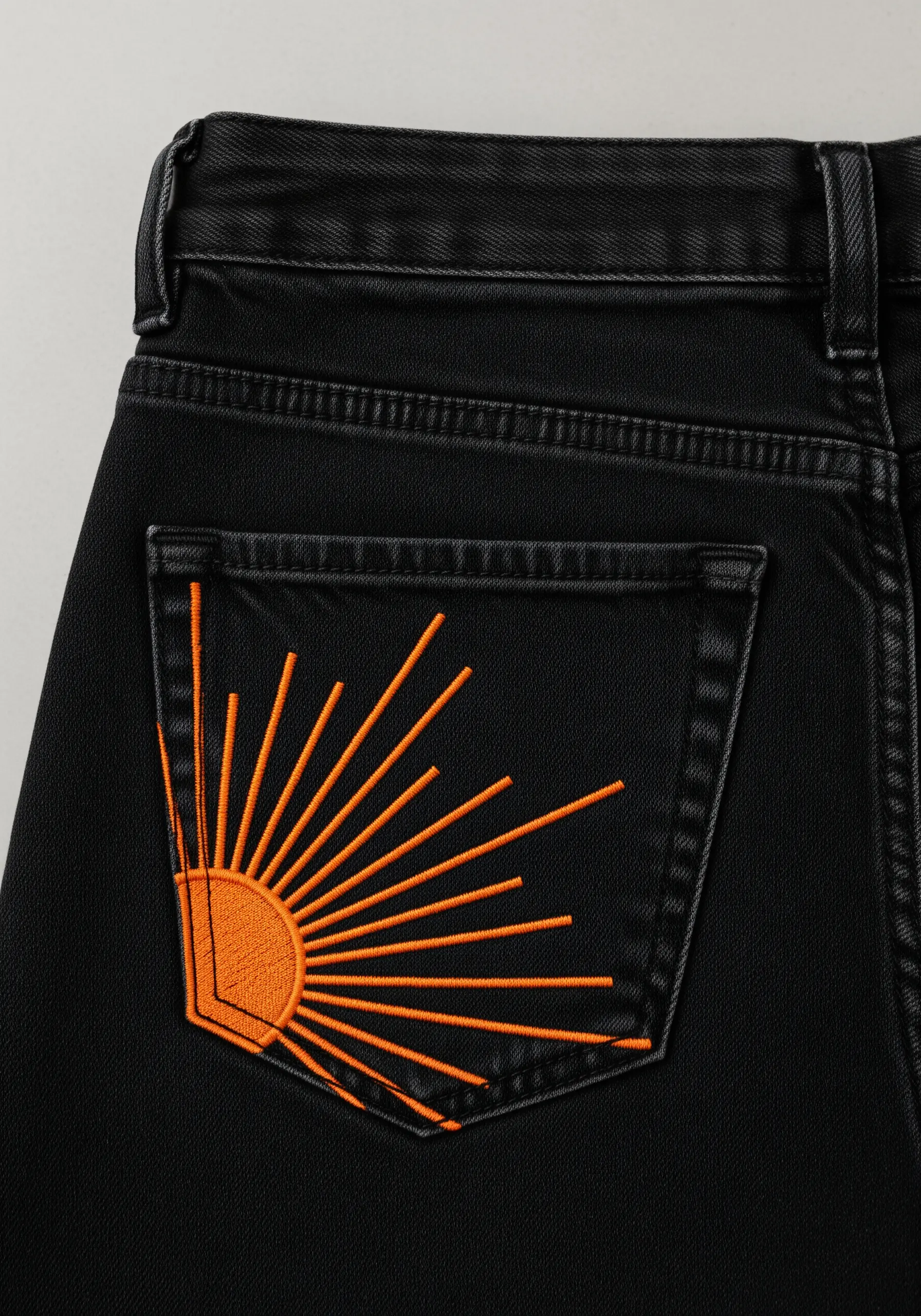 Bright orange sunburst graphic embroidered on the back pocket of black jeans.