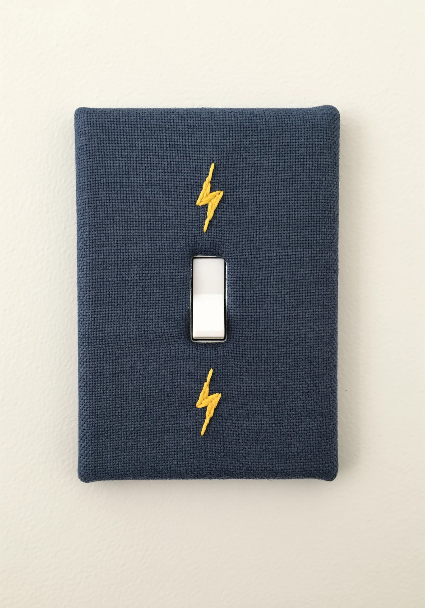 Two small yellow lightning bolts embroidered on a navy fabric light switch cover.