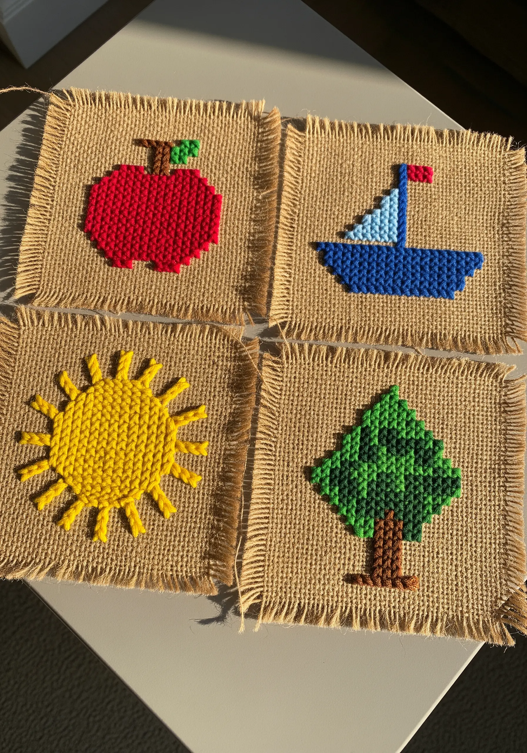 Four burlap squares with needlepoint designs: an apple, boat, sun, and tree.