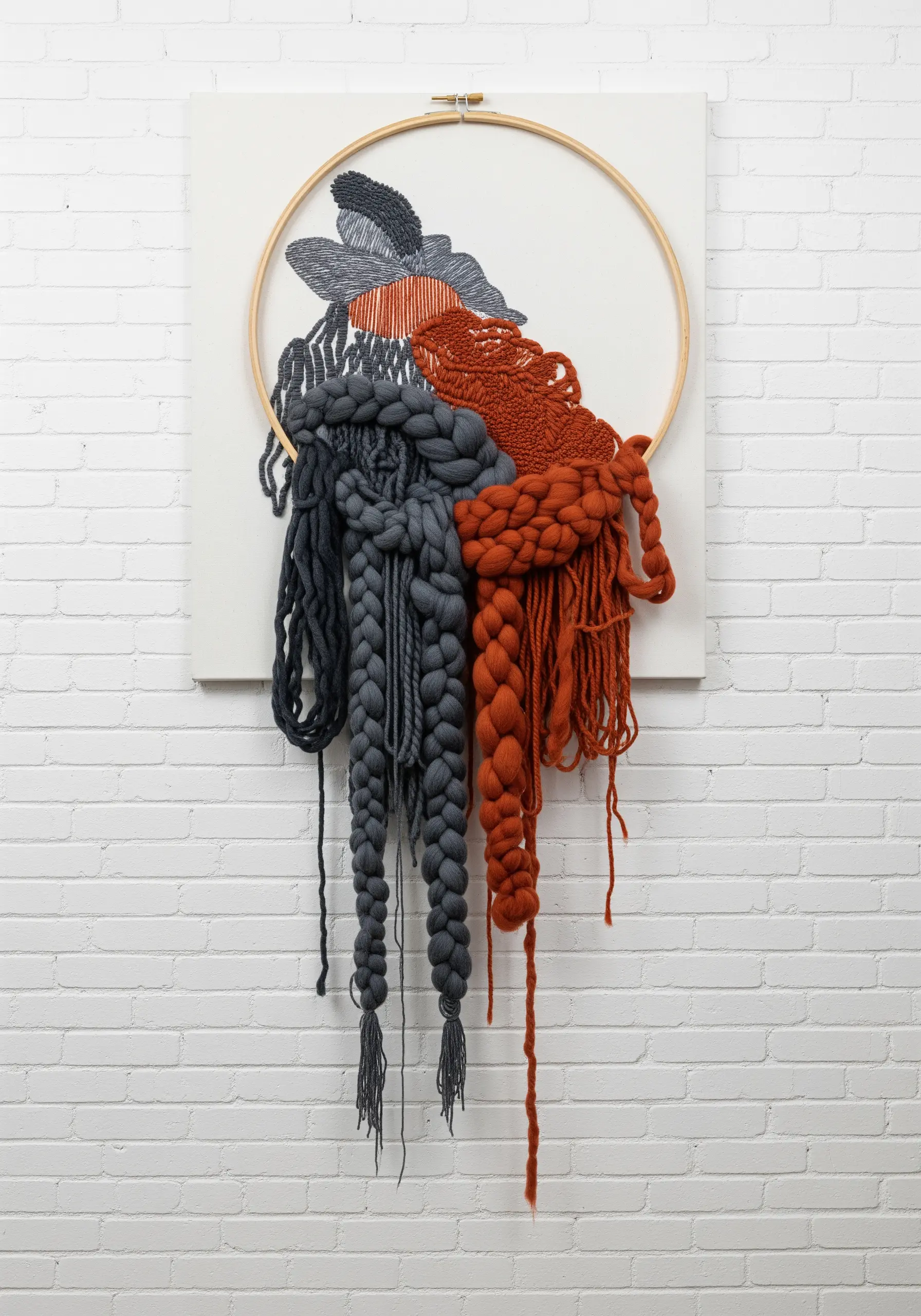 A mixed-media piece where oversized grey and rust-colored braids spill out of an embroidery hoop.