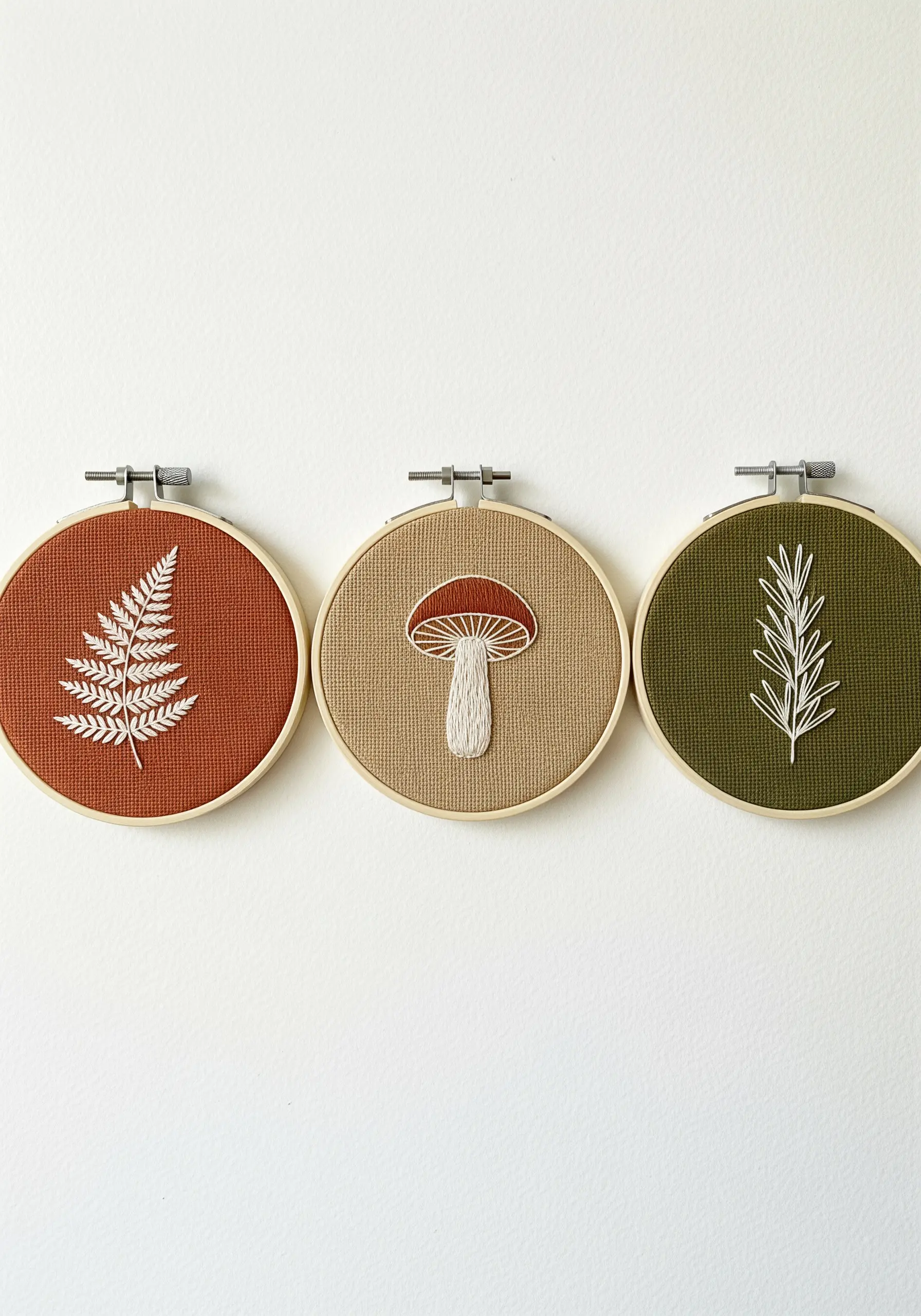 A trio of small embroidery hoops with fern, mushroom, and rosemary motifs on colored fabric.