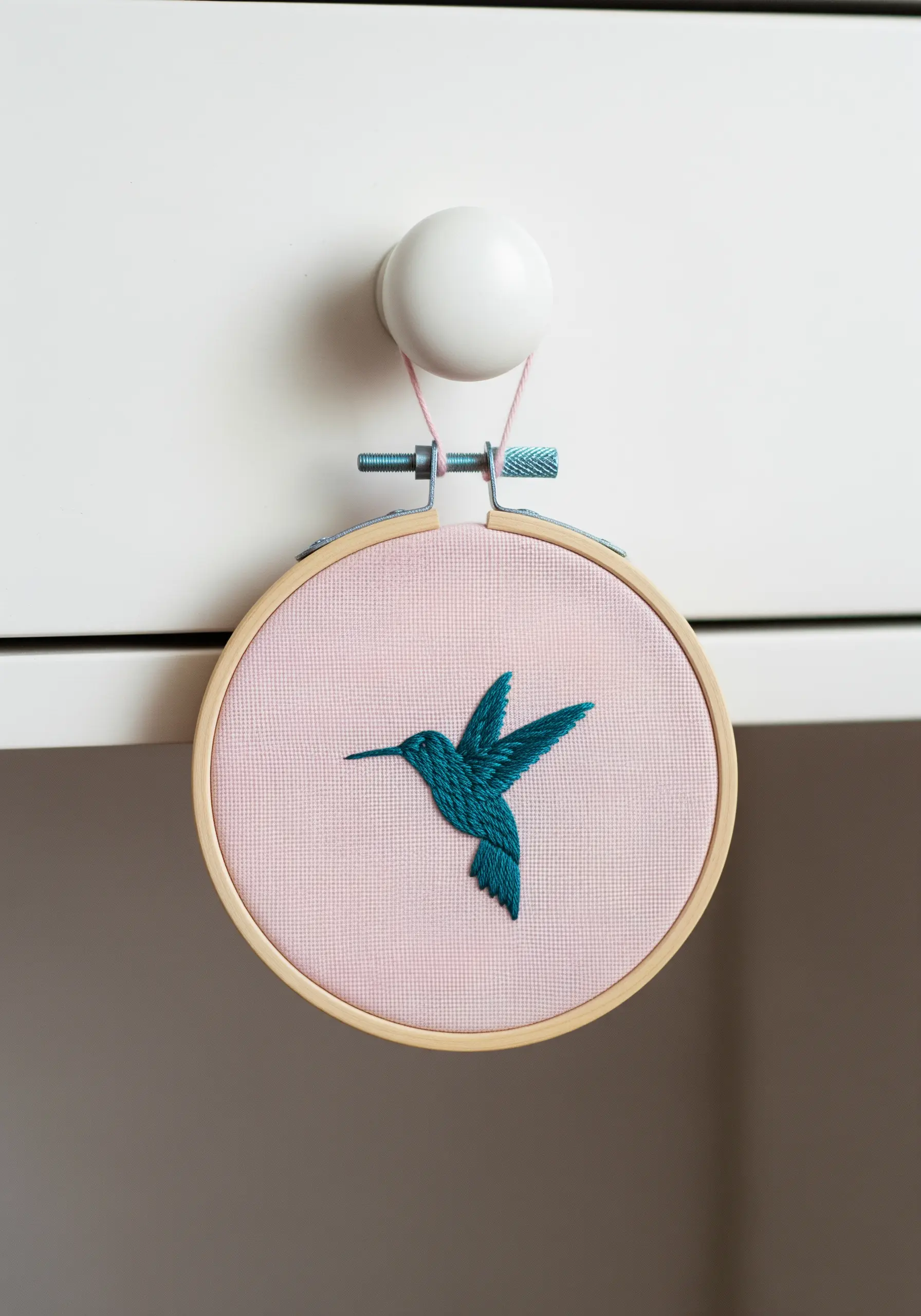 A small embroidery hoop with a turquoise hummingbird hanging from a white drawer knob.