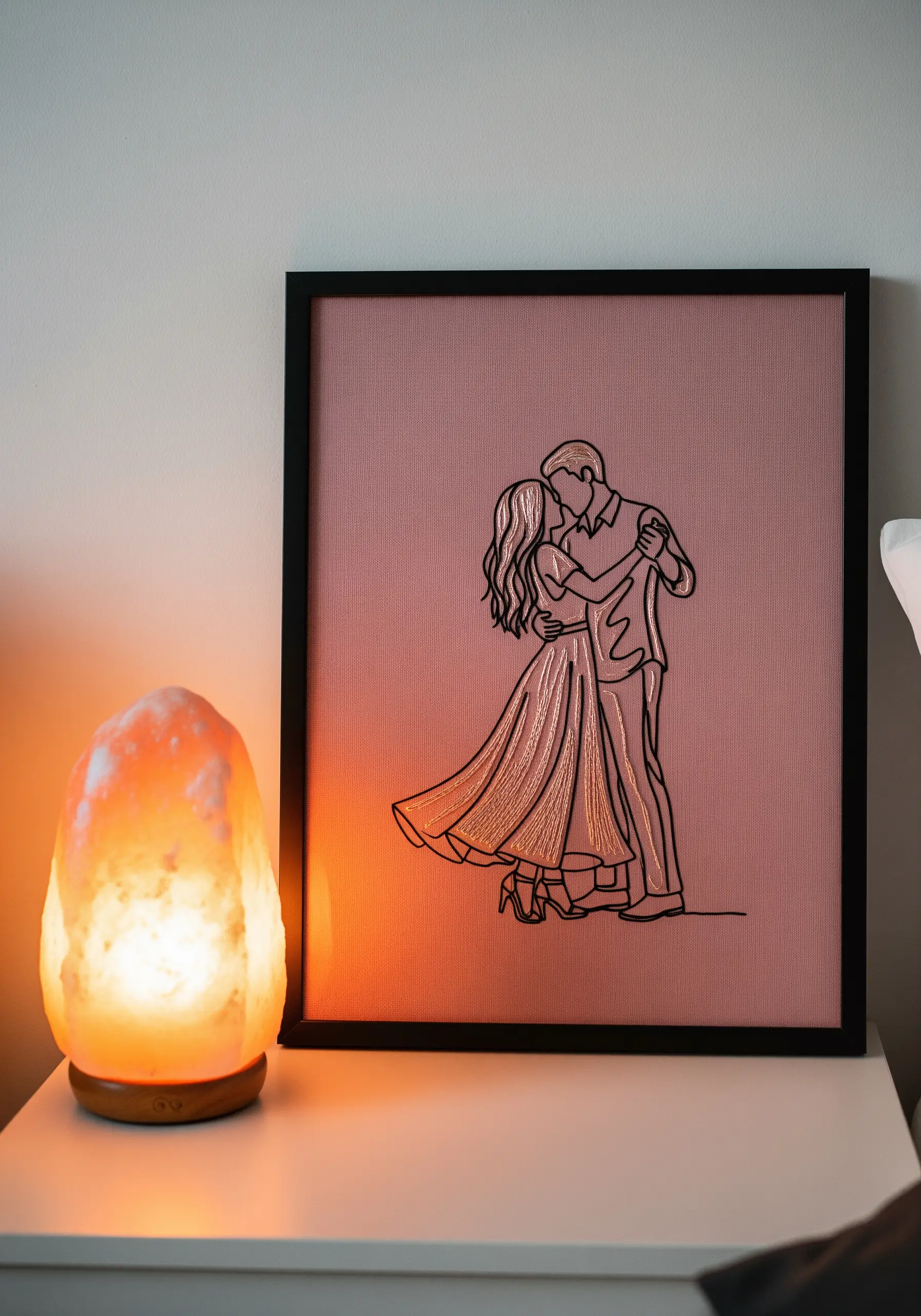 Framed embroidery of a dancing couple on dusty rose fabric, with white thread fill.