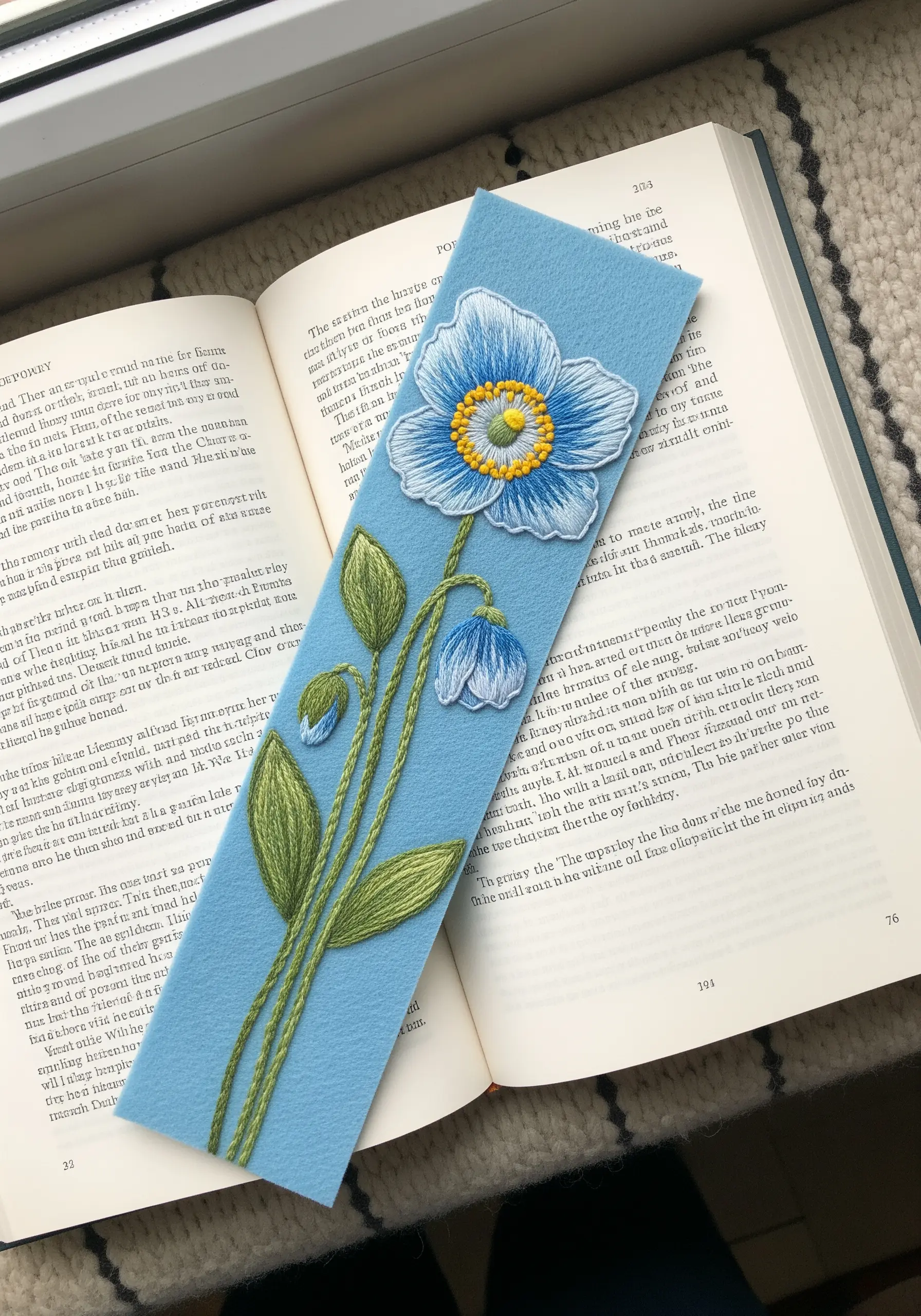 A hand-embroidered blue poppy on a light blue felt bookmark resting on an open book.