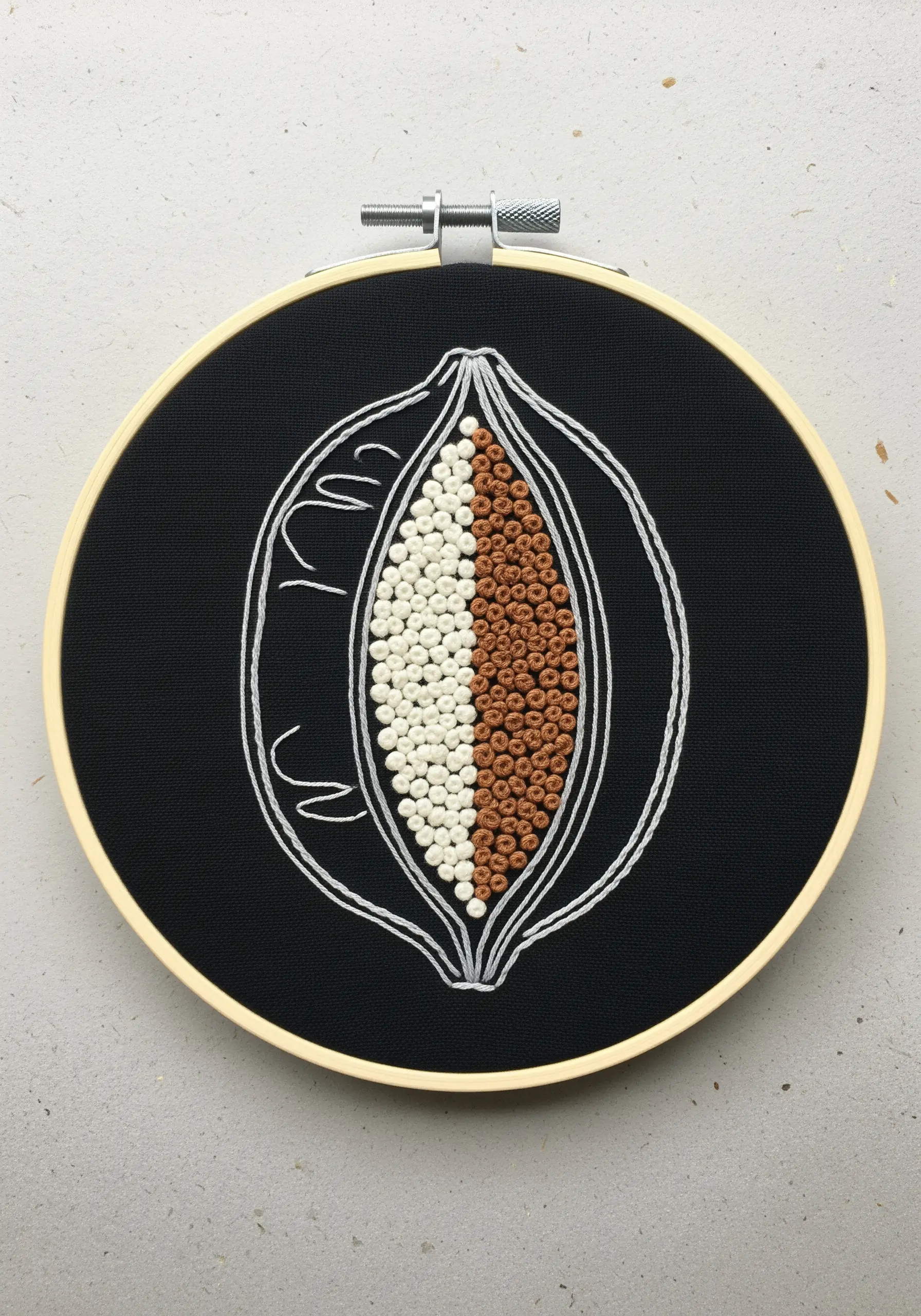 Abstract embroidery of a cacao pod filled with white and brown French knots on black fabric.