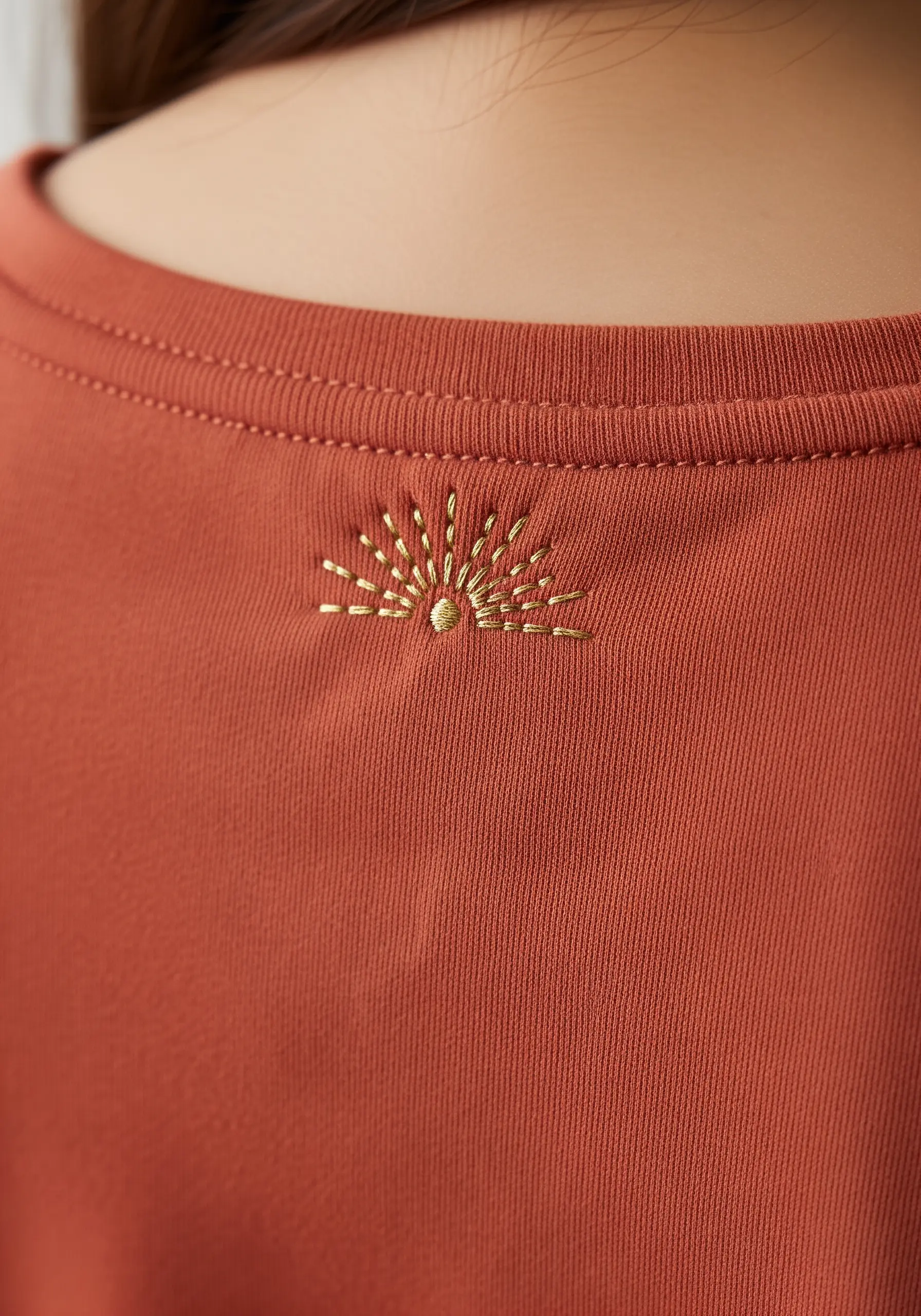 A minimalist sunrise embroidered with radiating gold metallic thread on a rust-colored t-shirt.