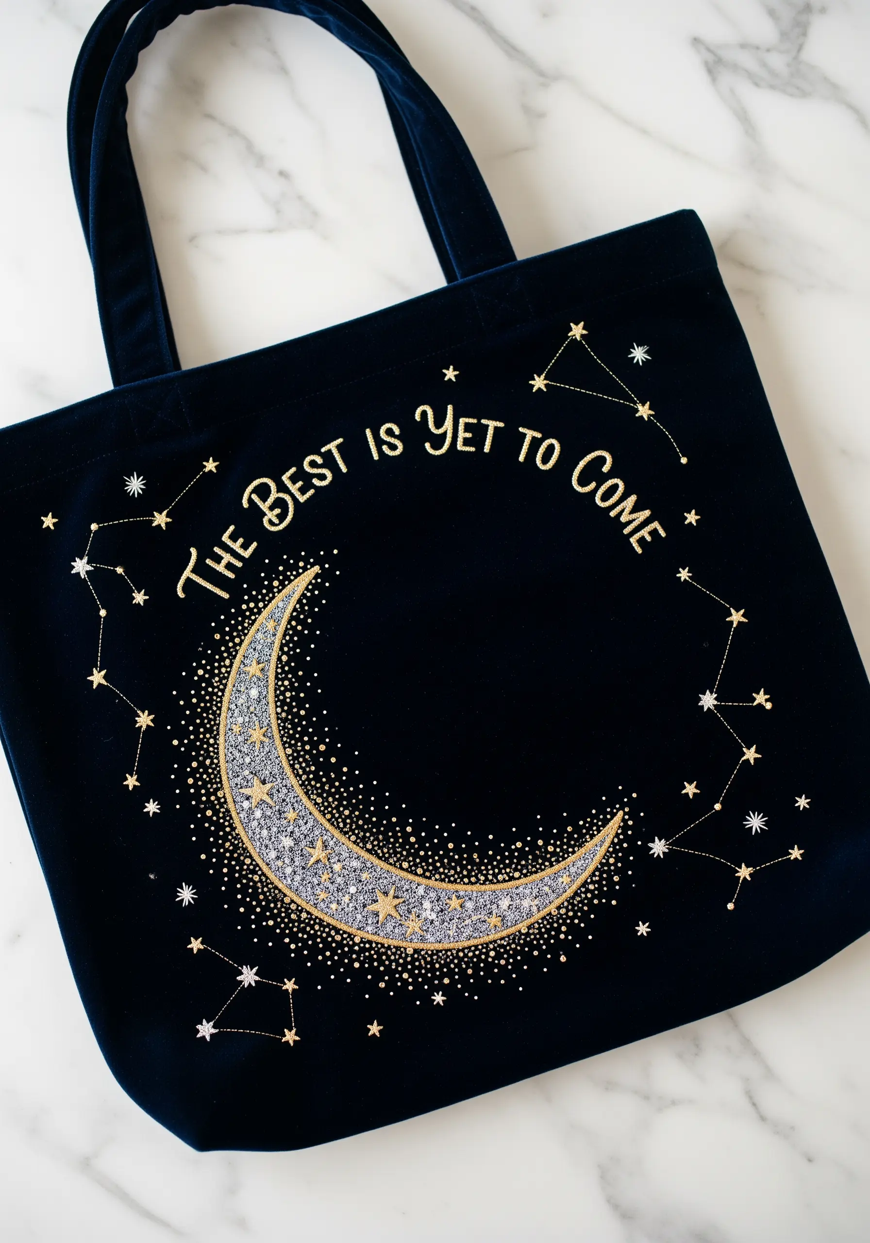 A crescent moon and constellations embroidered in metallic thread on a dark blue velvet tote bag.