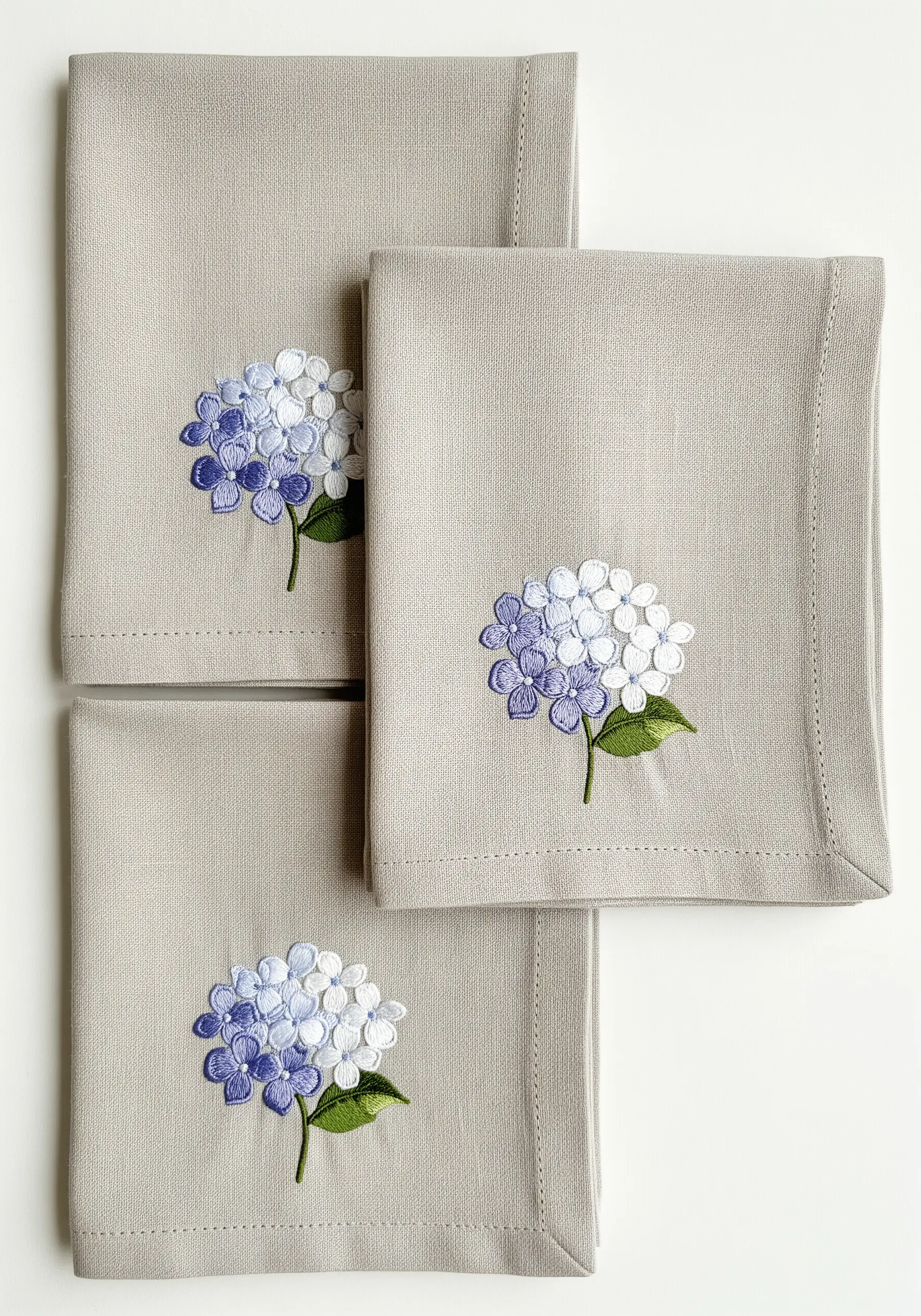 Three linen napkins, each with a small, minimalist blue and white hydrangea motif.