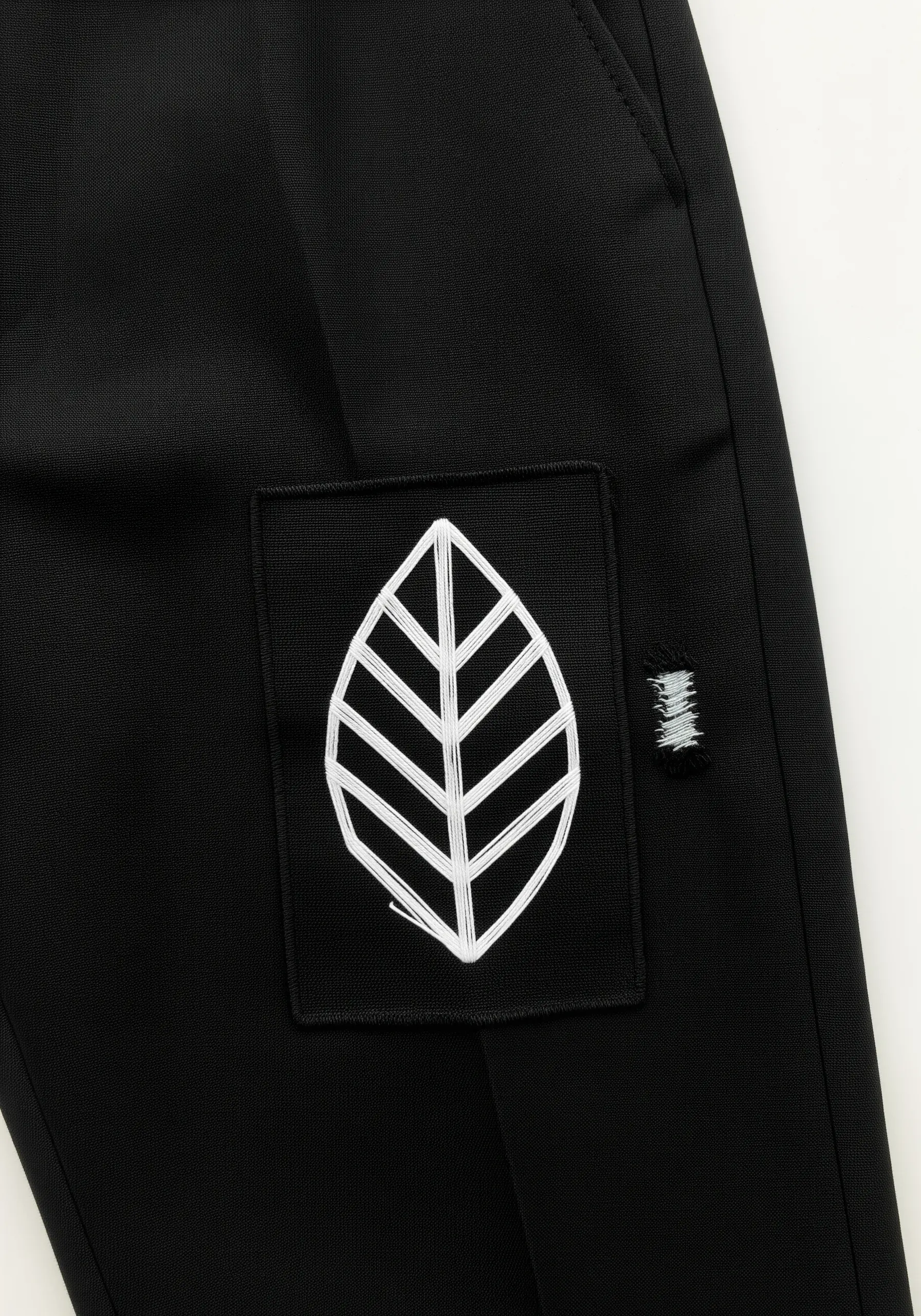 A minimalist black patch on black pants with a leaf motif stitched in thick white thread.